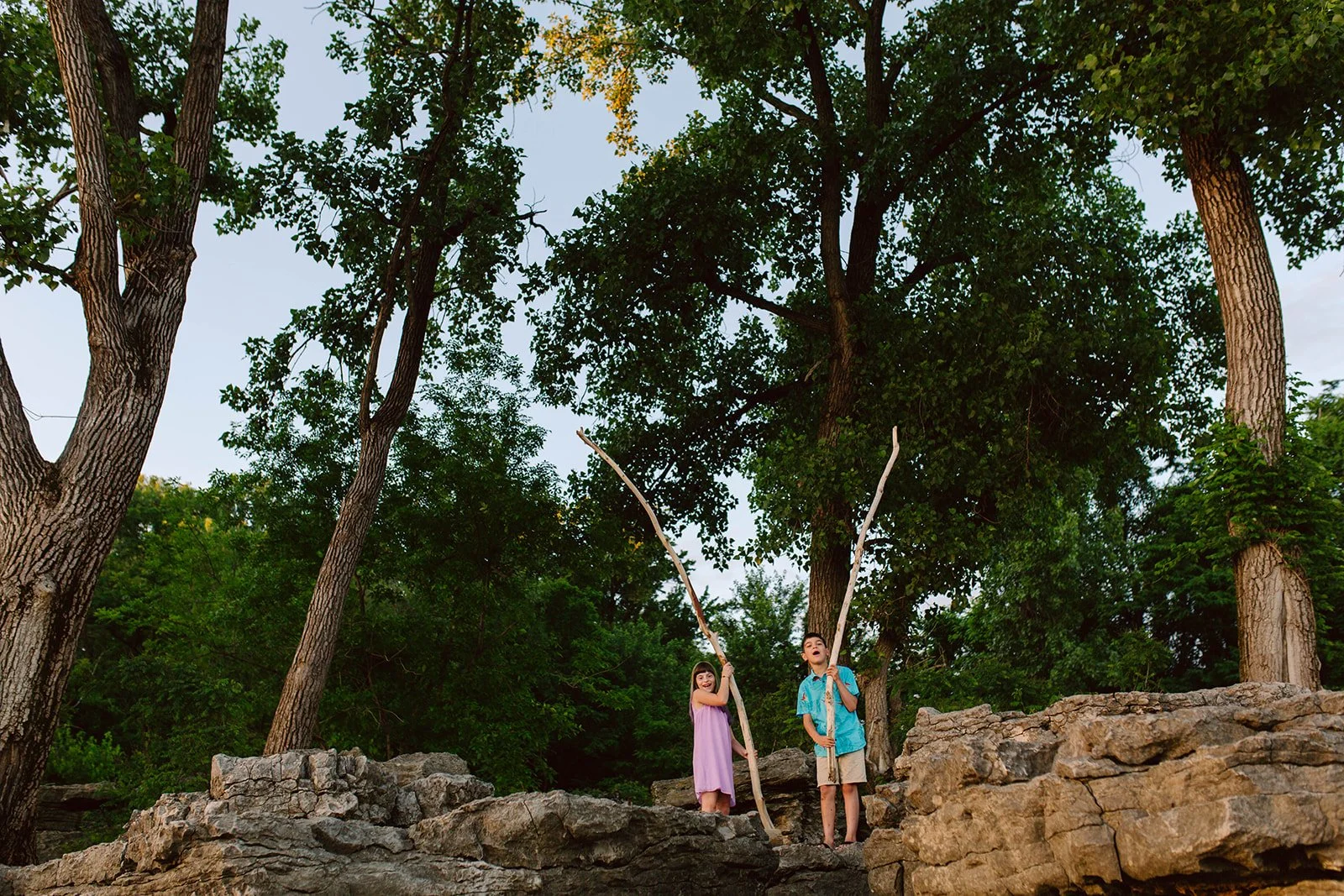 Two children, a girl in a purple dress and a boy in a blue shirt and shorts, standing on rocks in a wooded area, holding long sticks.