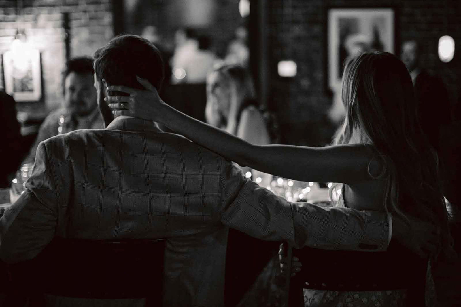 A woman with long hair is touching the neck of a man wearing a patterned blazer at a dinner gathering. The setting appears to be a dimly lit restaurant with other guests in the background.