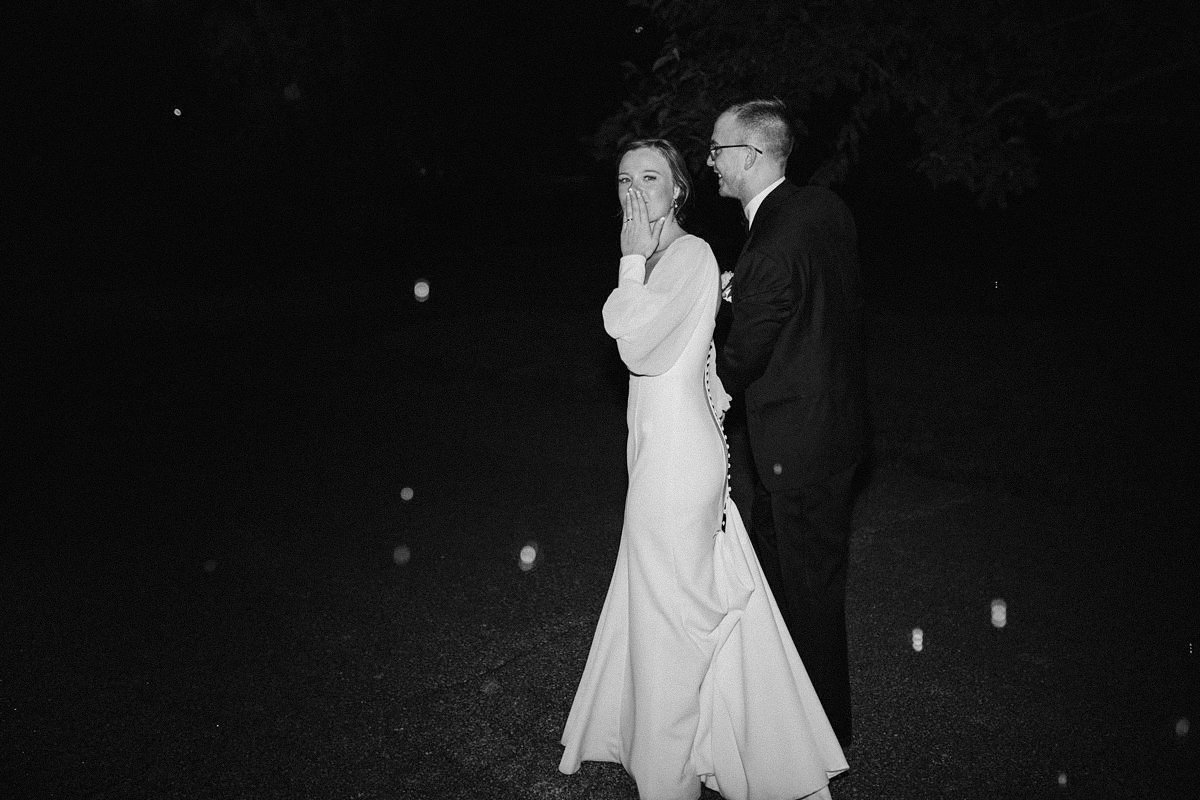 A bride in a long white dress covering her mouth with her hand, standing next to a groom in a suit, at night outdoors.