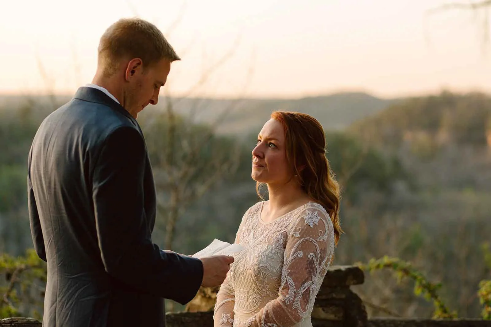 A man and woman exchanging vows outdoors at sunset, with the man reading from a paper and the woman looking at him lovingly.