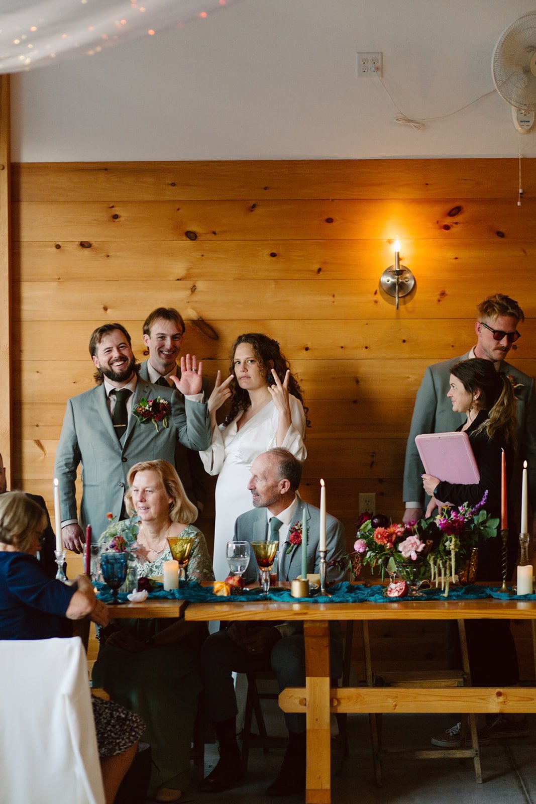 Group of people celebrating at a wedding reception with a wooden wall background, decorated table, and candles.