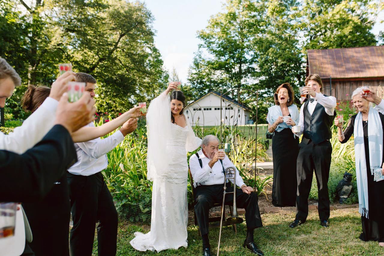 A group of people celebrating outdoors with drinks during a wedding, featuring a bride in a white dress and an elderly man with a cane raising a glass.