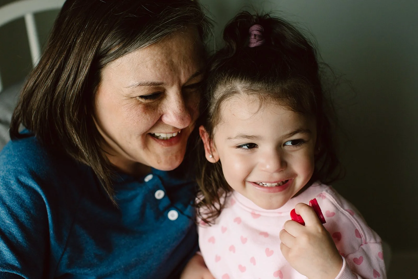 A woman and a young girl smiling and close together indoors, with the woman leaning her head against the girl, who is holding a red toy or object.