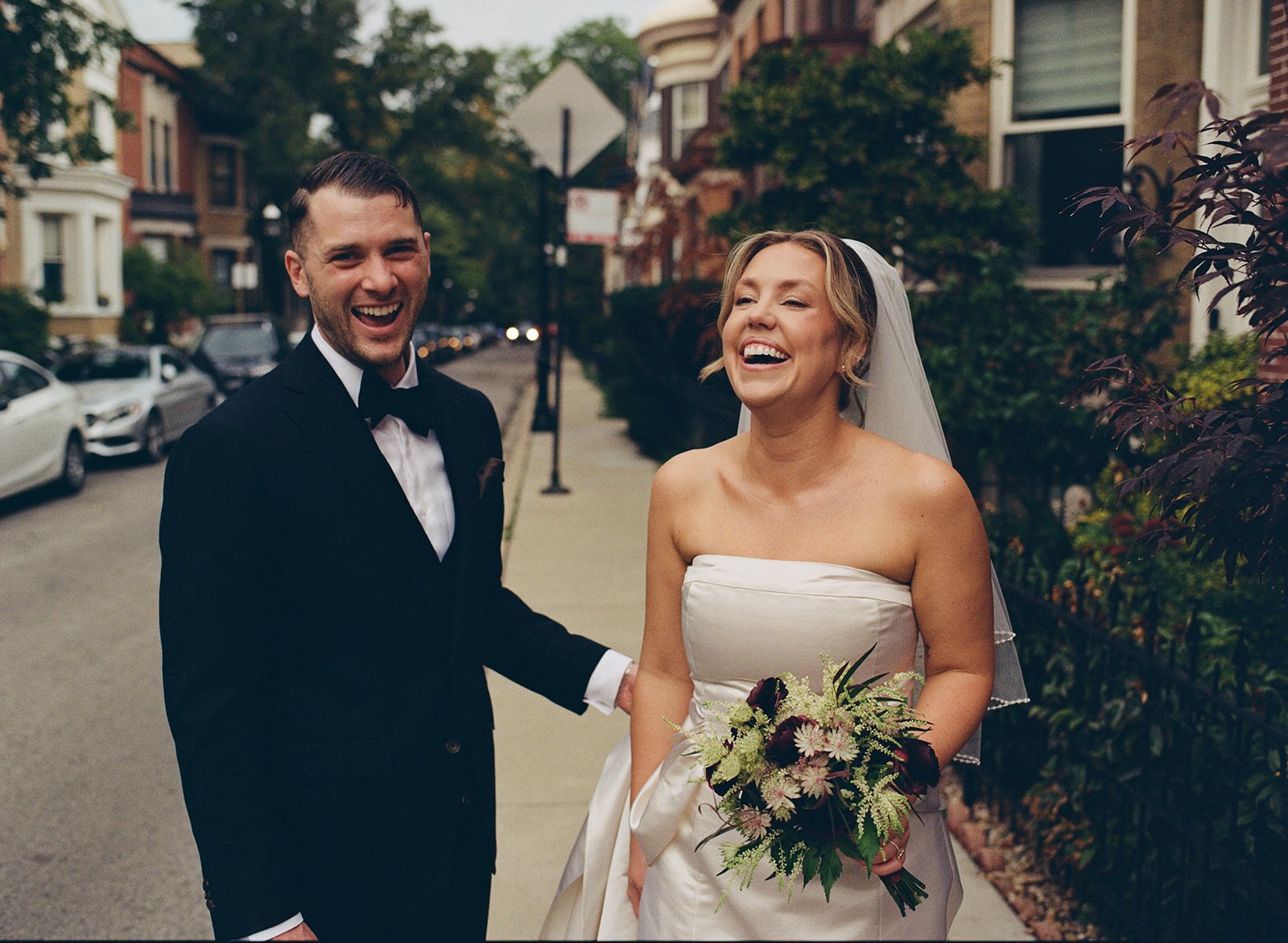 A smiling bride and groom laughing on a tree-lined city street, with parked cars and historic houses in the background. The bride is holding a bouquet of flowers and wearing a strapless wedding dress and veil. The groom is in a black tuxedo with a bow tie.