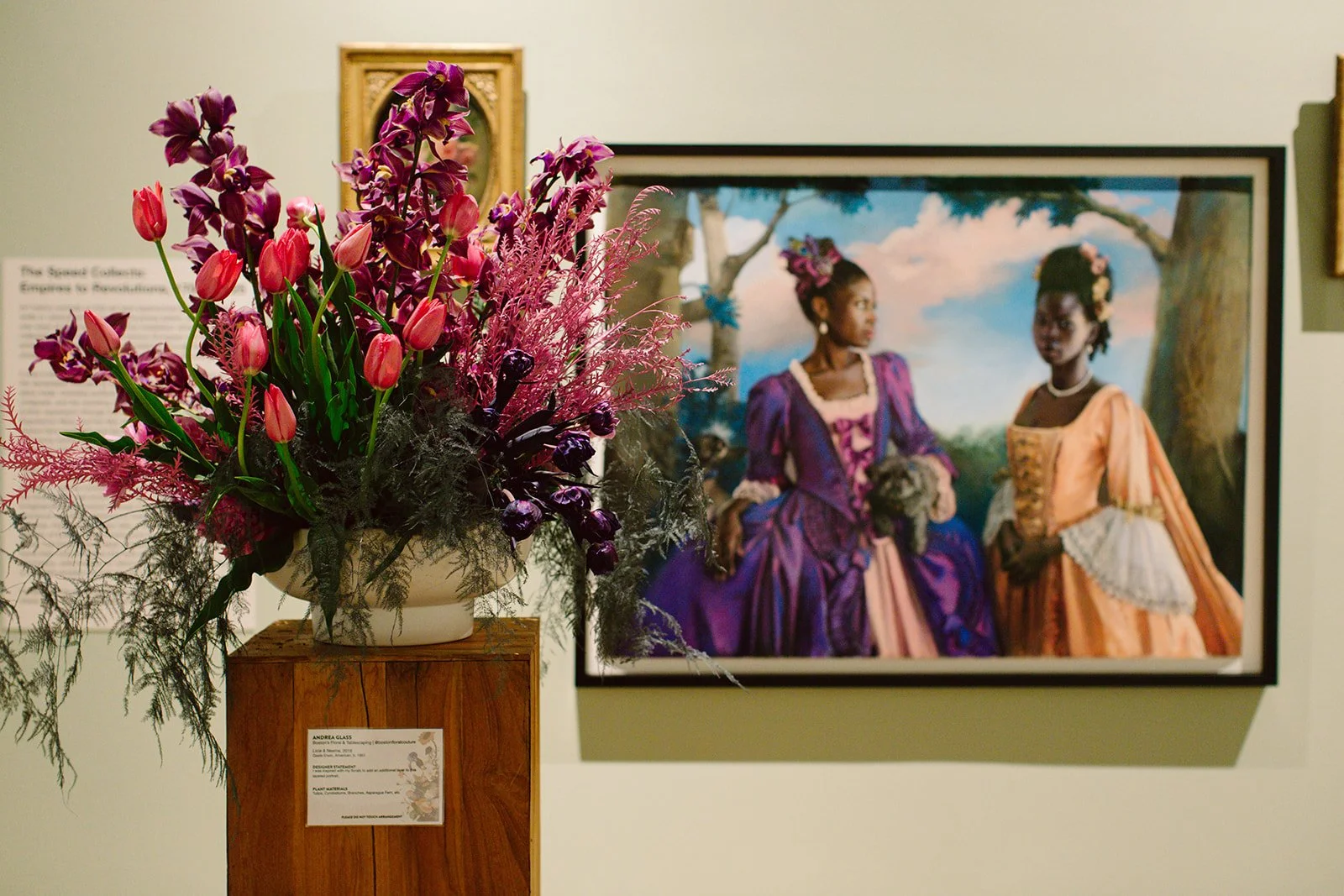 A floral arrangement of pink tulips, purple orchids, and pink feathers in a white vase on a wooden pedestal in an art gallery with a painting of two women in historical attire hanging on the wall behind.