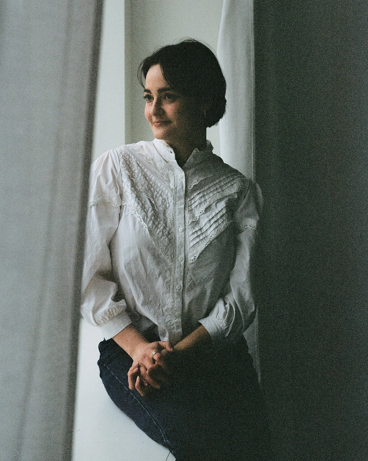 Woman with short dark hair wearing a white embroidered blouse and dark jeans, standing indoors near a window with her hands clasped.