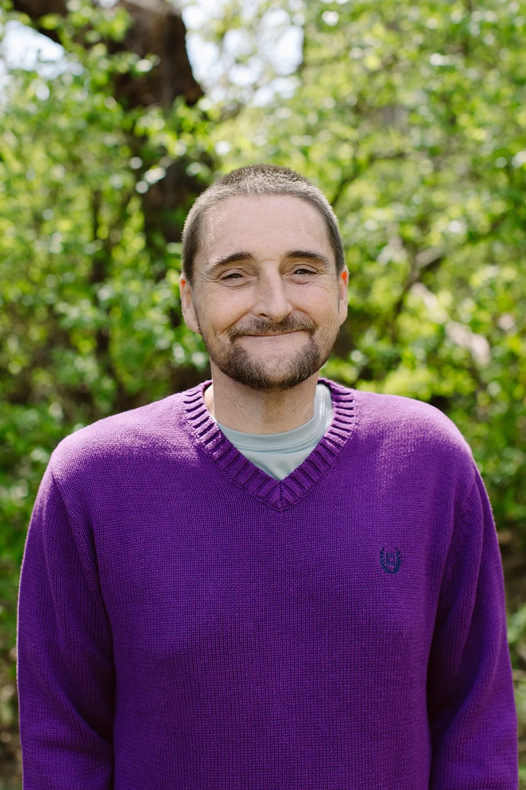 A man smiling outdoors, wearing a purple sweater over a white shirt, with a green leafy background.