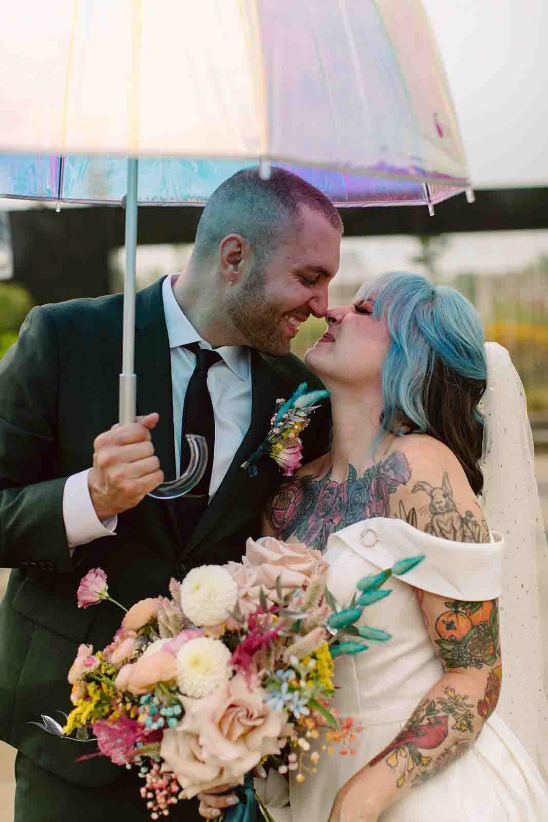 A couple, one with blue hair and tattoos, and the other in a black suit, sharing a moment under a rainbow umbrella during their wedding, both smiling warmly.