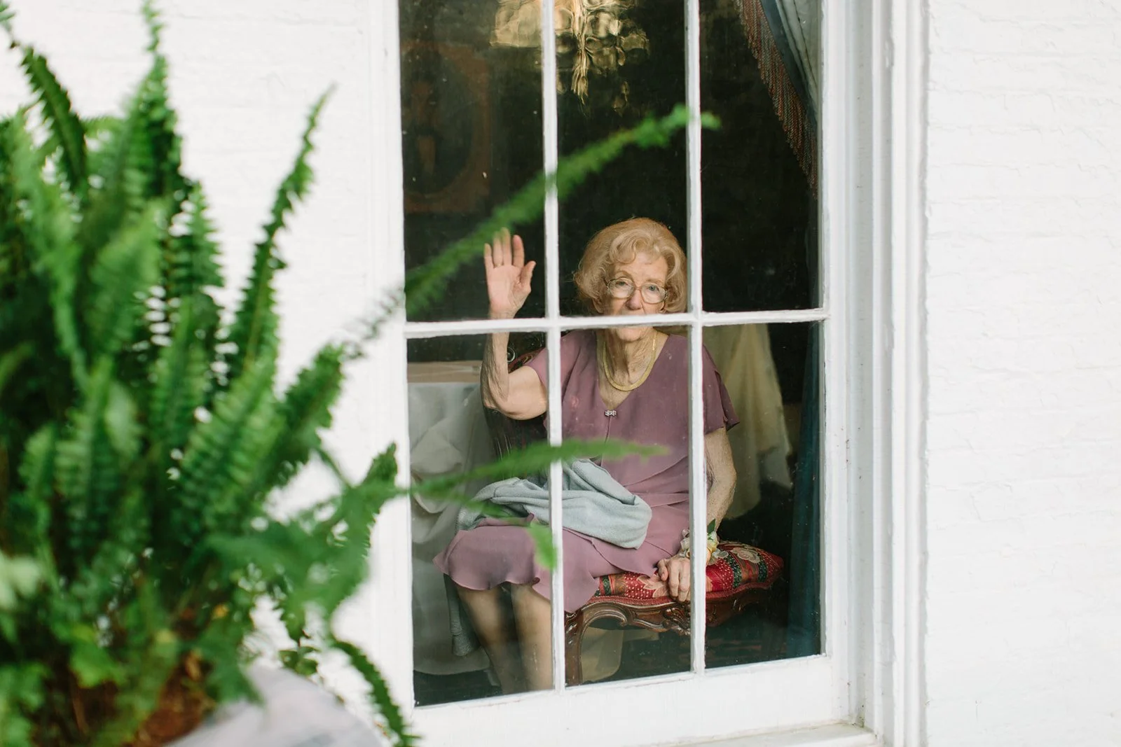 An elderly woman with glasses, sitting inside a house behind a window, waving her hand and smiling.