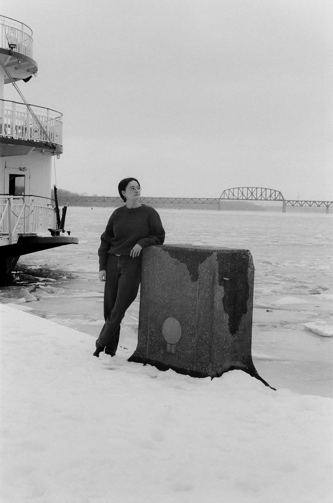 A woman standing on a snow-covered shore, leaning against a concrete barrier near the water. A boat is docked nearby, and a bridge is visible in the distance across the water.