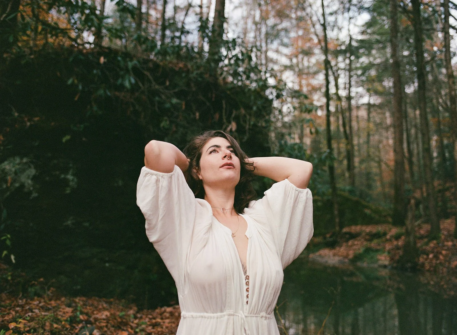 A woman with dark brown hair and wearing a white dress with puffy sleeves standing outdoors in a forest with tall trees and autumn leaves, looking up with her hands behind her head.