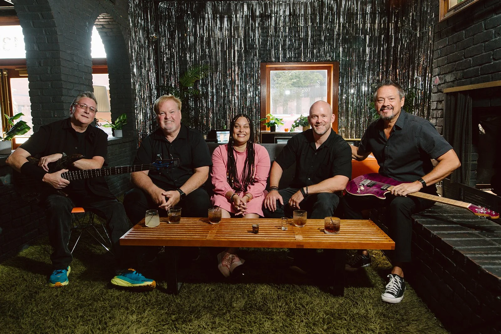 A group of five people sitting around a low wooden table in a cozy, dimly lit room. Two of them are holding electric guitars. There are drinks on the table, and the background features dark brick walls and decorative metallic curtains.