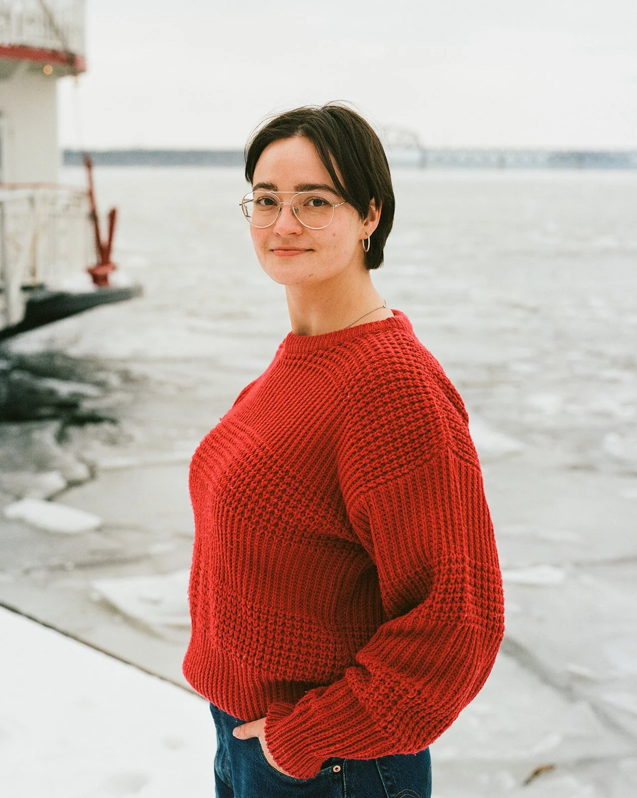A woman standing outdoors near a body of water wearing a red sweater and glasses.
