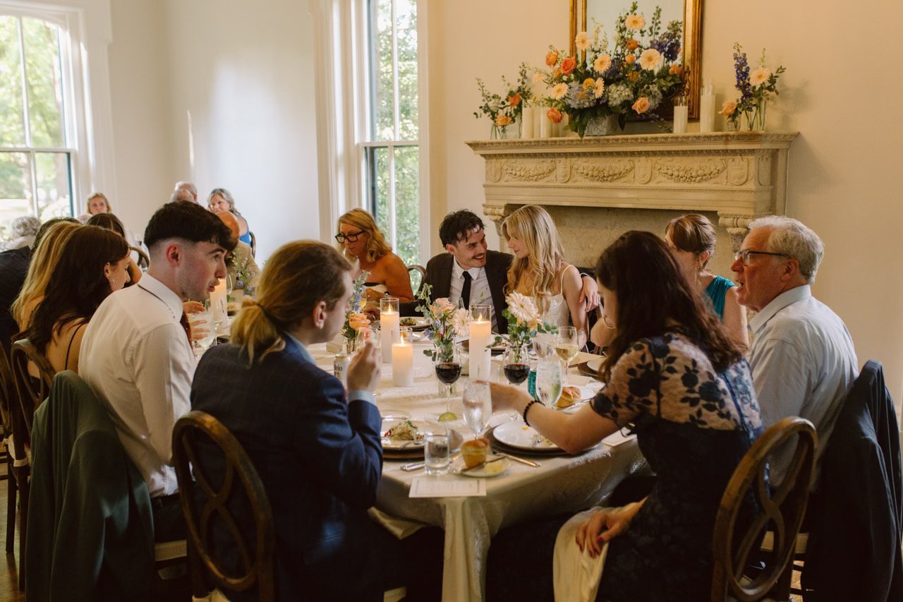 People sitting around a banquet table during a wedding reception, with floral arrangements, lit candles, and food, in a well-lit room with large windows and a decorative fireplace with floral decorations above it.
