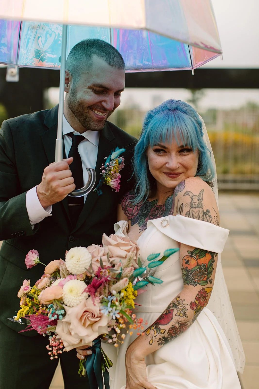 bride and groom pose under a colorful umbrella while holding a bouquet of flowers
