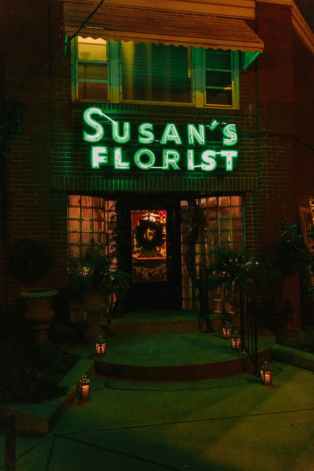 Nighttime exterior view of Susan's Florist with a green neon sign. The door is decorated with a wreath, and there are potted plants. The building has brick walls.