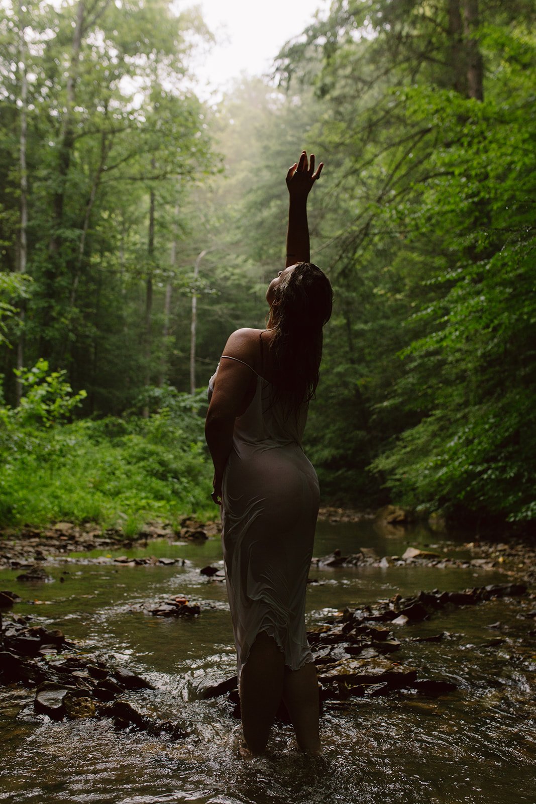 A woman in a dress standing in a creek in a forest, reaching upward with one hand, surrounded by green trees.