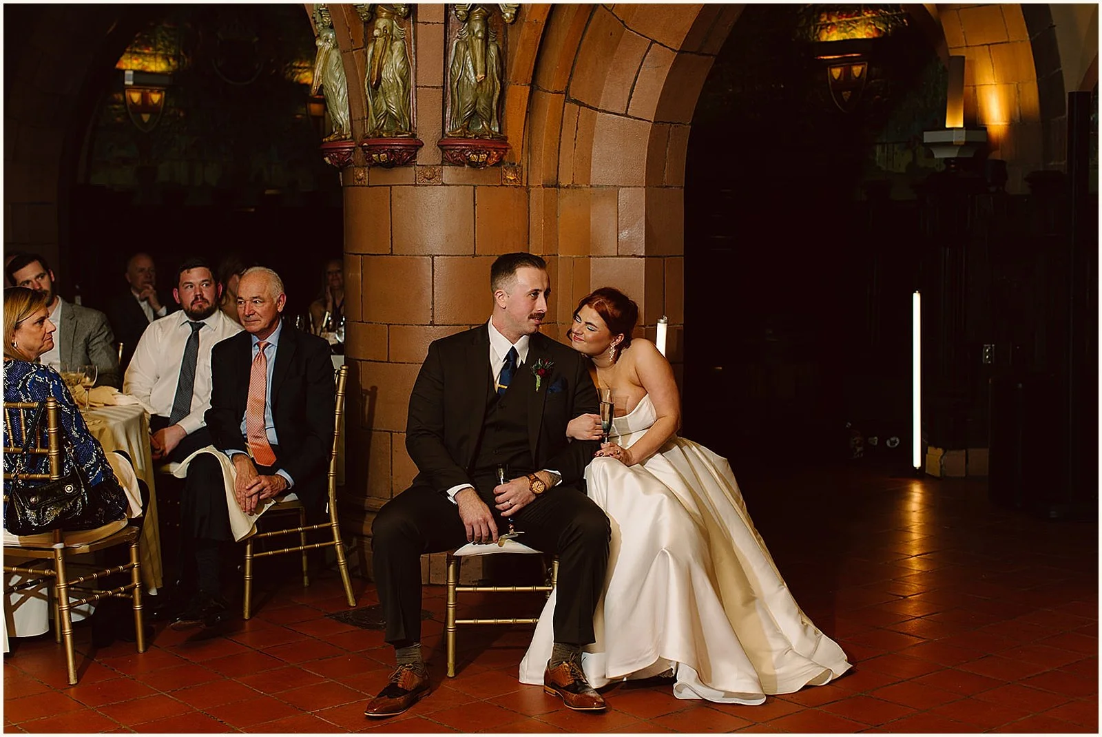 A bride and groom sit on chairs listening to a speech.