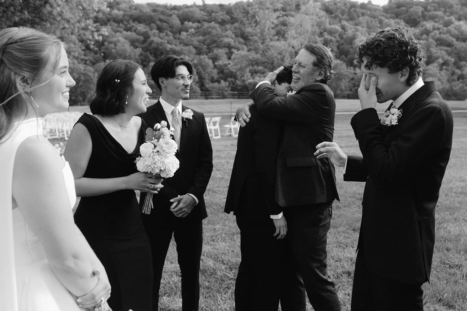 A black-and-white photo of a wedding scene with five people outdoors, including a bride in a white dress, a woman holding a bouquet, a man in a suit, and two men in suits, one of whom appears emotional, with trees and outdoor chairs in the background.