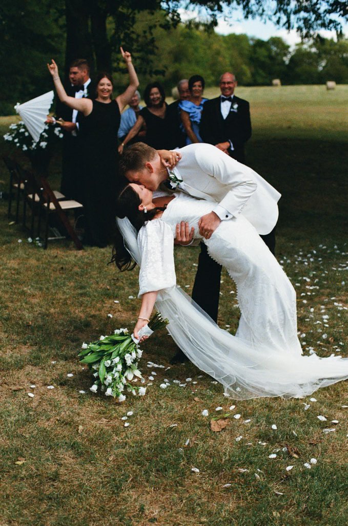 A bride and groom share a kiss after their wedding ceremony outdoors, with friends and family in the background.