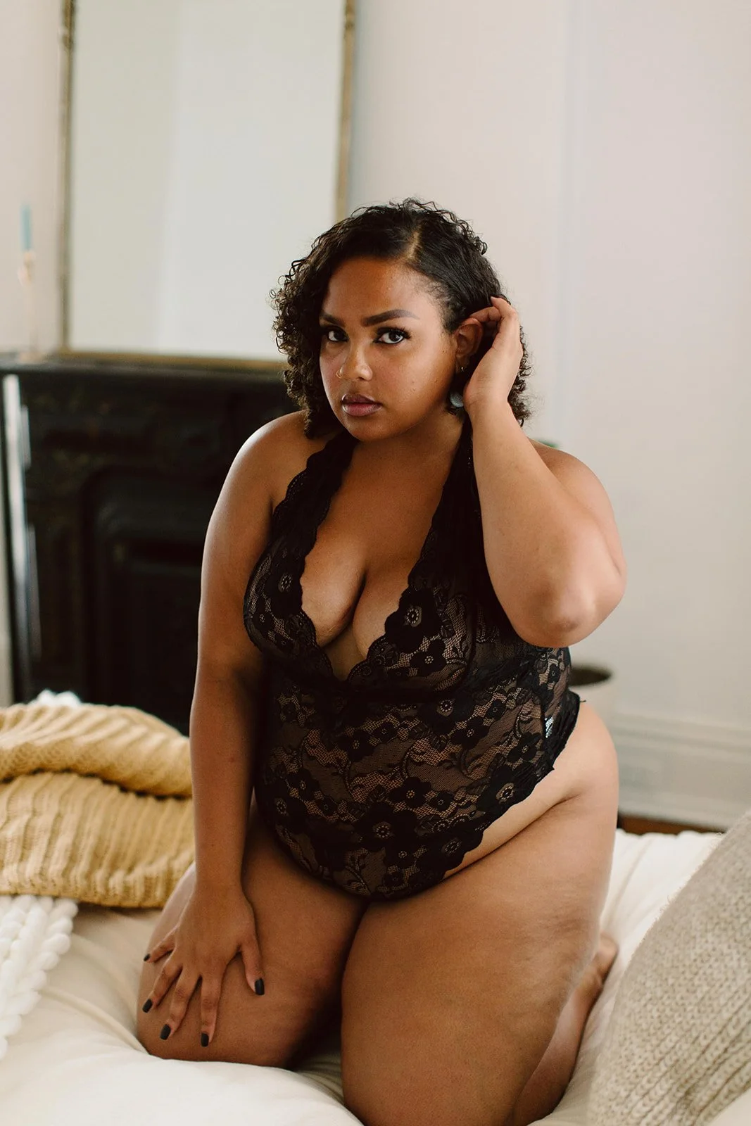 A woman with dark curly hair in black lace lingerie sitting on a bed with beige and white blankets, touching her hair, in a softly lit room.