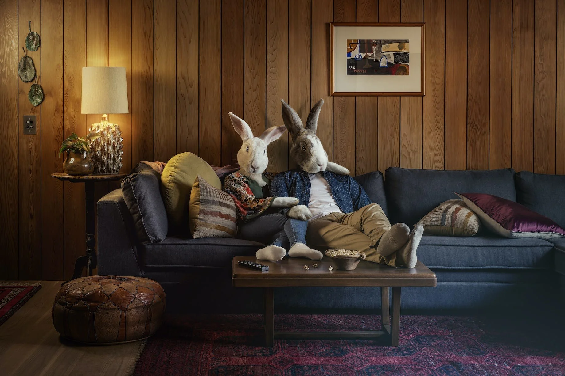 Two people with rabbit heads sitting on a dark blue sofa in a wood-paneled living room. One person wears a colorful scarf, and the other wears a blue shirt. There are pillows on the sofa, and a coffee table in front with snacks and a remote control. 