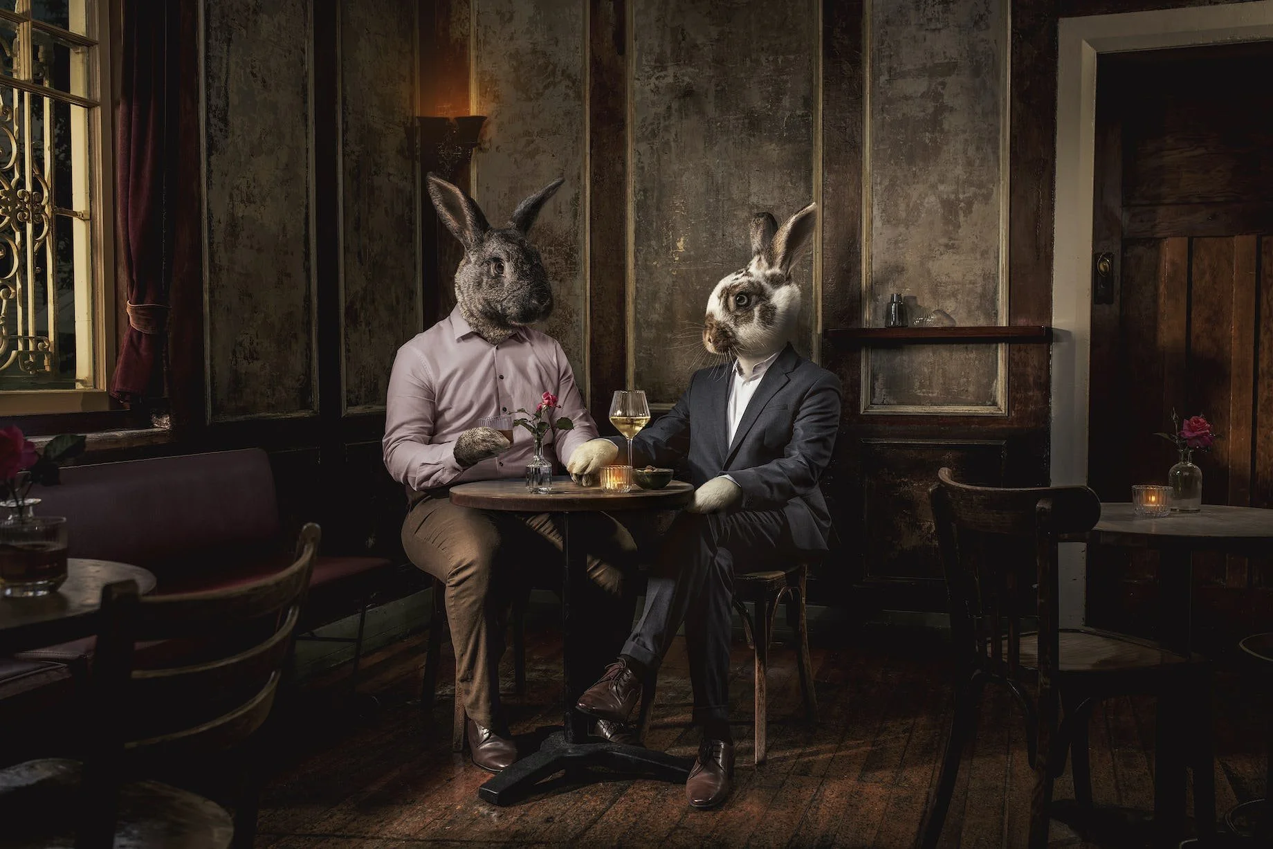 Two anthropomorphic rabbits with human bodies and rabbit heads sitting at a restaurant table. One rabbit is wearing a pink shirt, and the other is dressed in a suit. They are holding hands, with wine glasses and flowers on the table, in a cozy, dimly