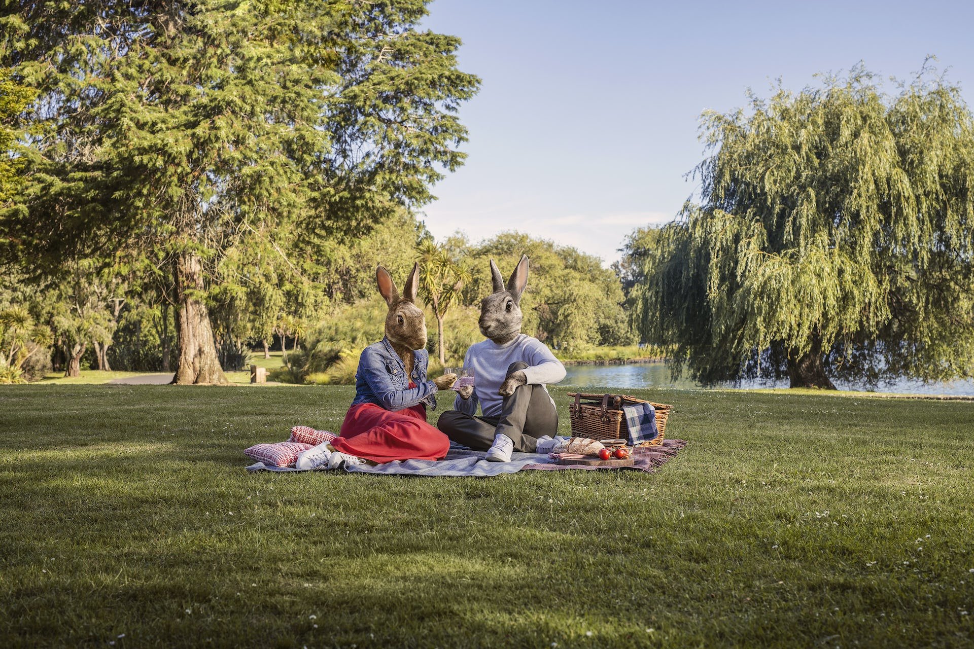Two rabbits with human bodies sitting on a blanket in a park having a picnic, surrounded by trees and a lake, with one rabbit holding a glass and the other reaching towards it.