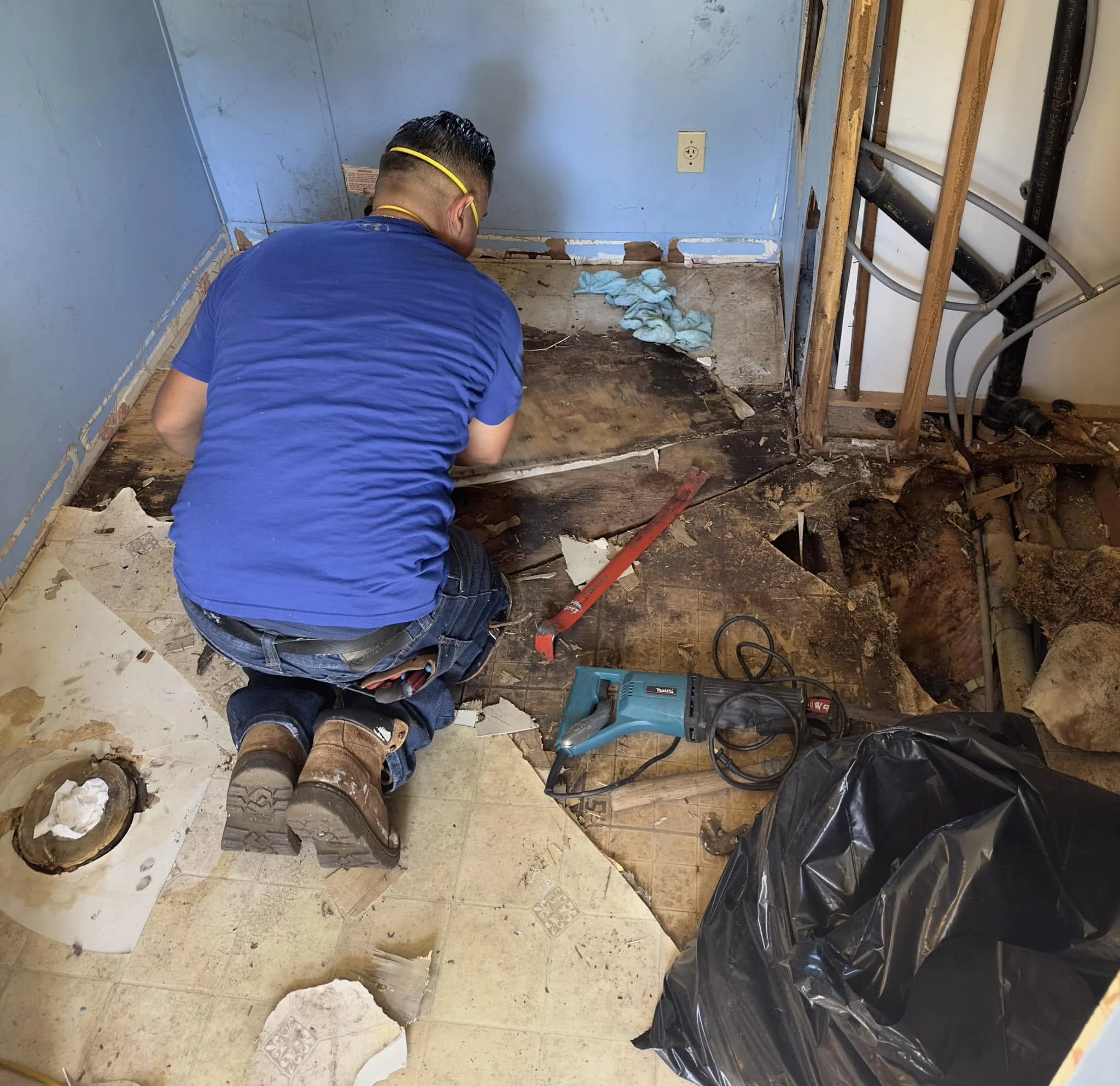 A person kneeling on the floor during a home renovation, removing old flooring with tools such as a pry bar and a power saw, with exposed subfloor and plumbing pipes visible.