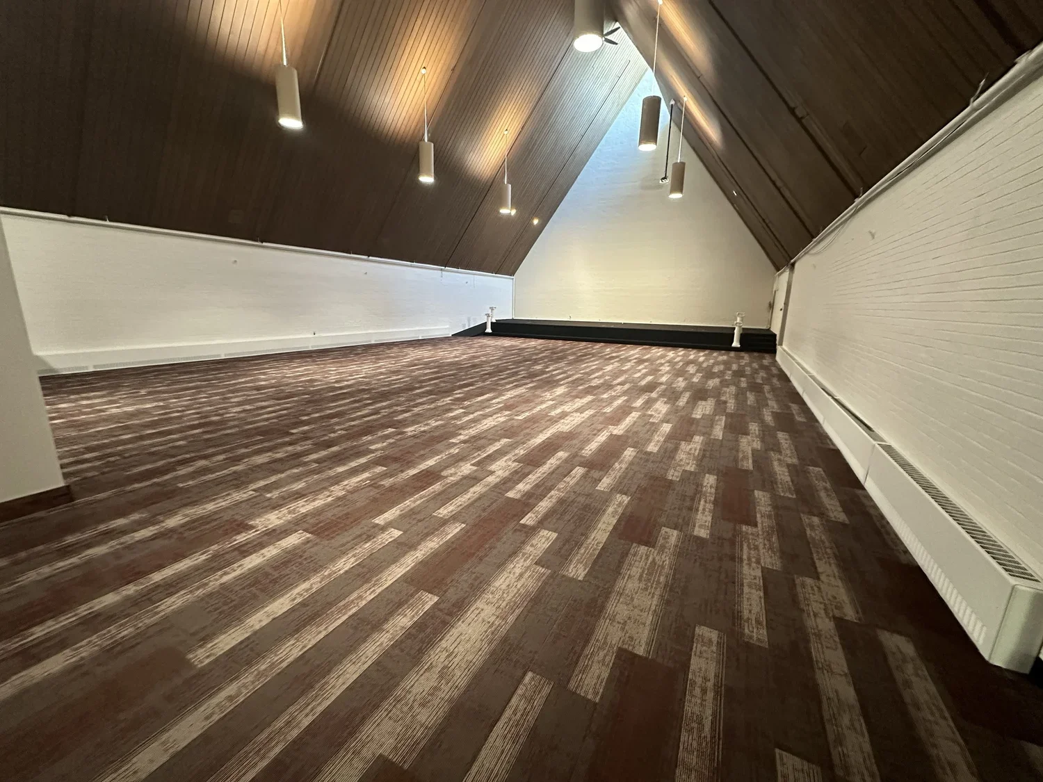 Empty attic room with sloped wooden ceiling, hanging pendant lights, white brick wall, patterned carpet, black baseboard, and a vent along the wall.