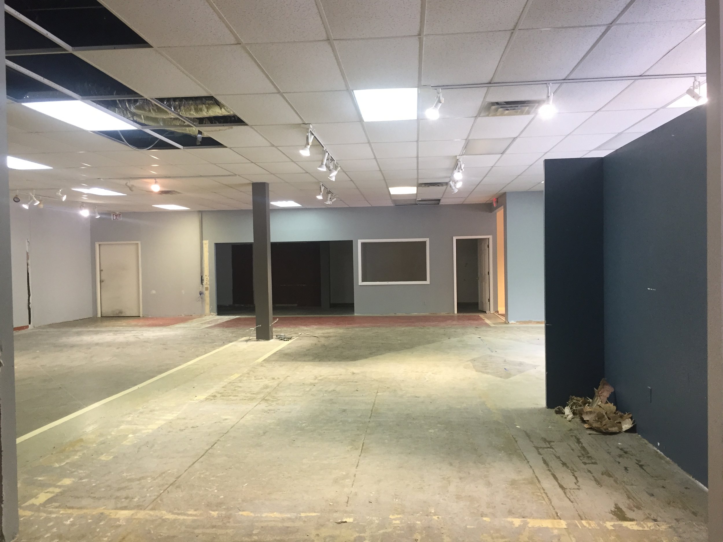 Empty commercial space undergoing renovation with exposed flooring, unfinished walls, and ceiling panels with some missing or damaged, and debris on the floor.