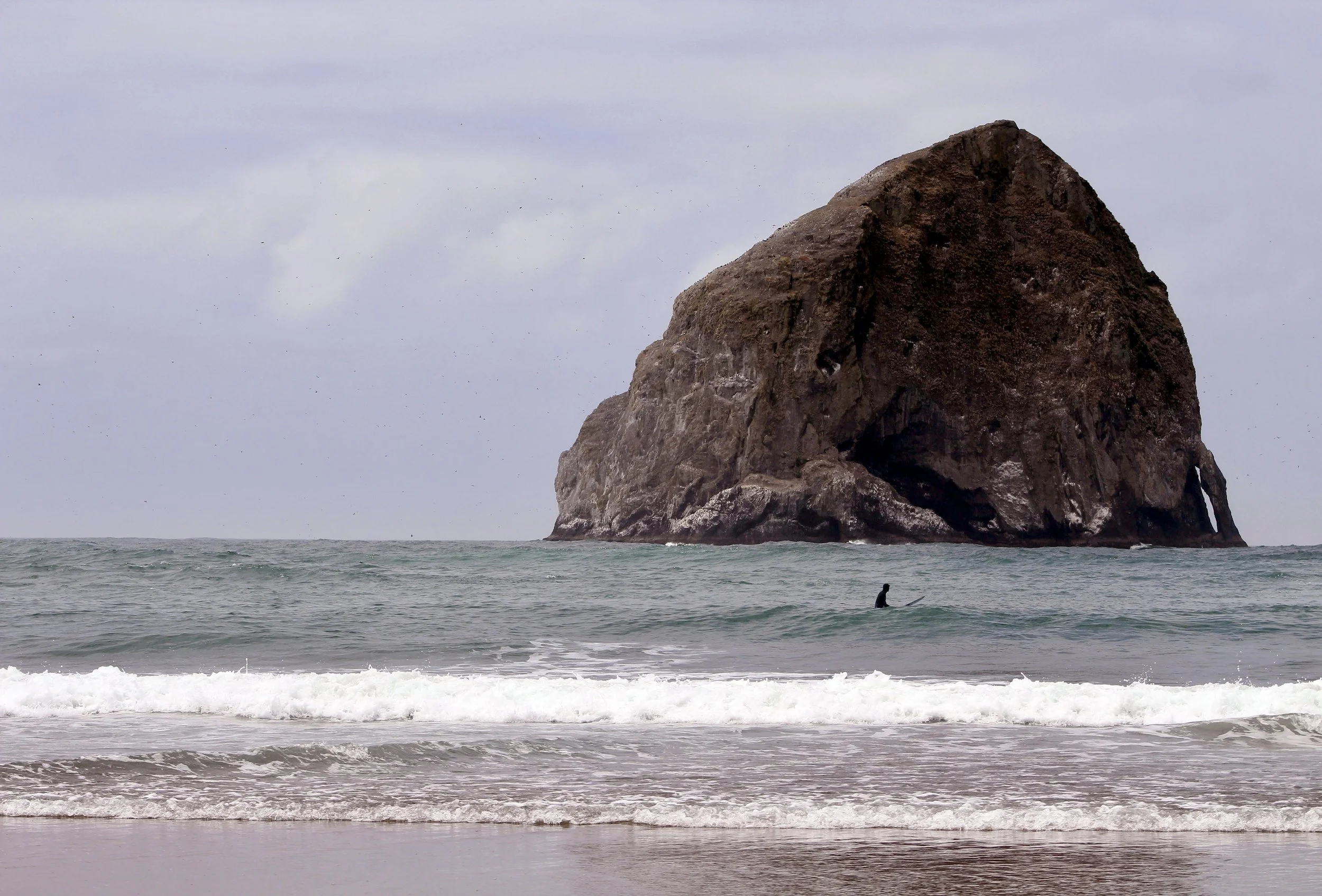 Surfer sitting on a board as waves break in front of a large rock formation rising from the ocean.