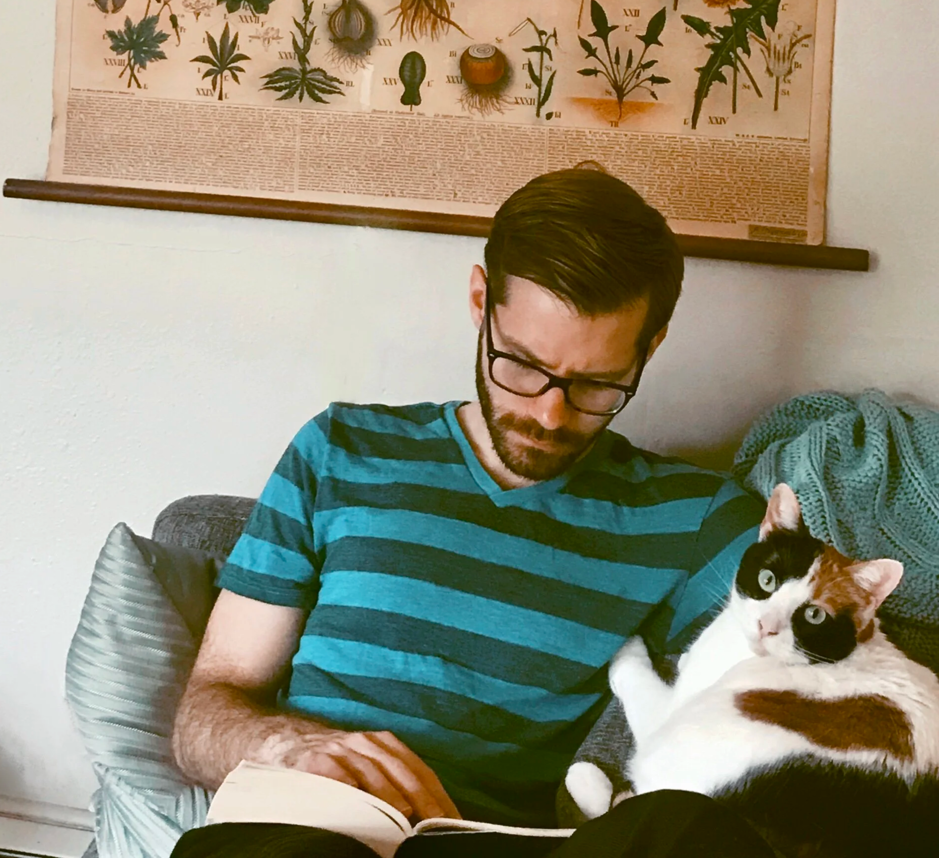 Ryan reading a book on a couch while a calico cat lays beside him.