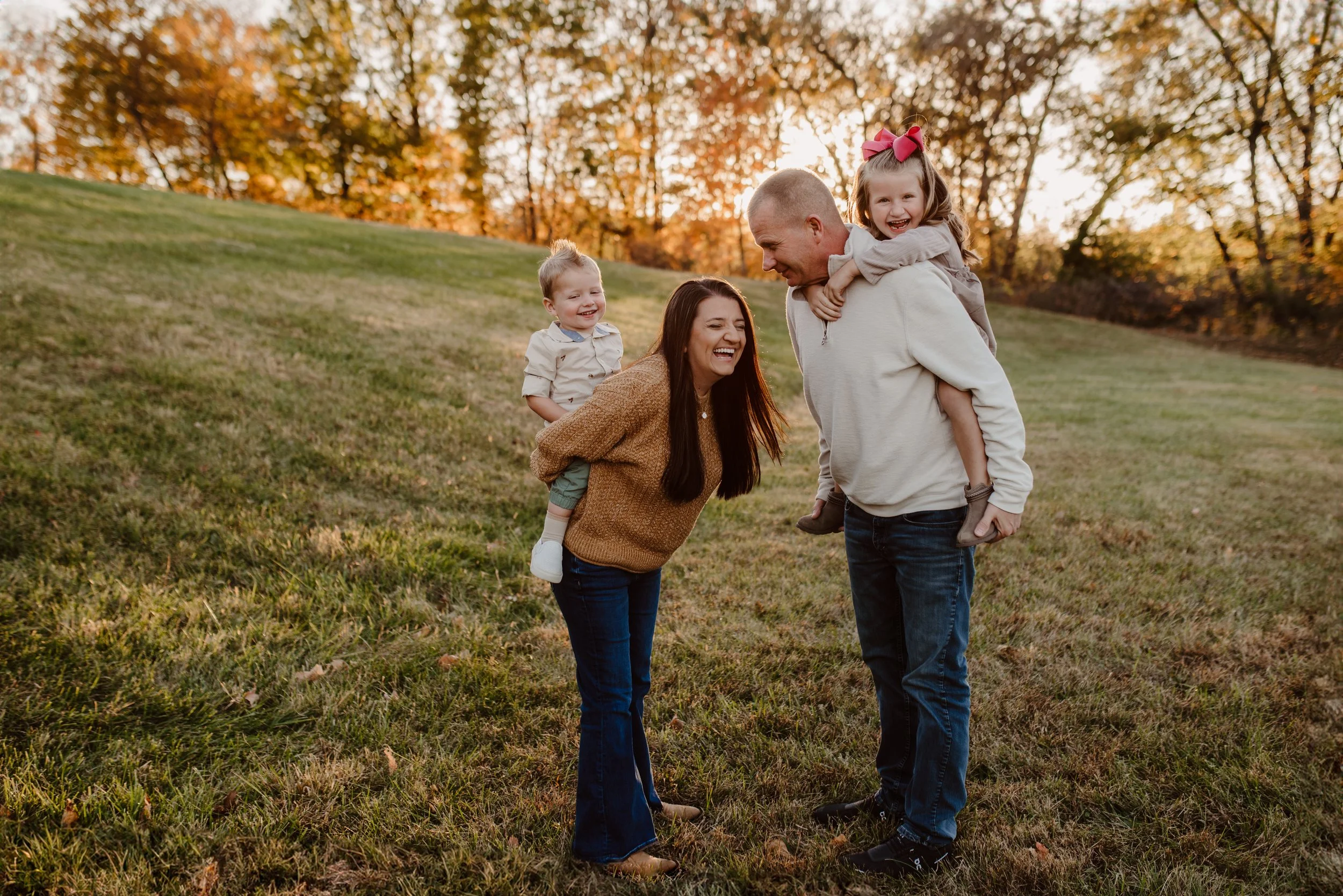 A family of four enjoying an outdoor moment in a grassy field during sunset; the father has a daughter on his back, while the mother and son stand and laugh.