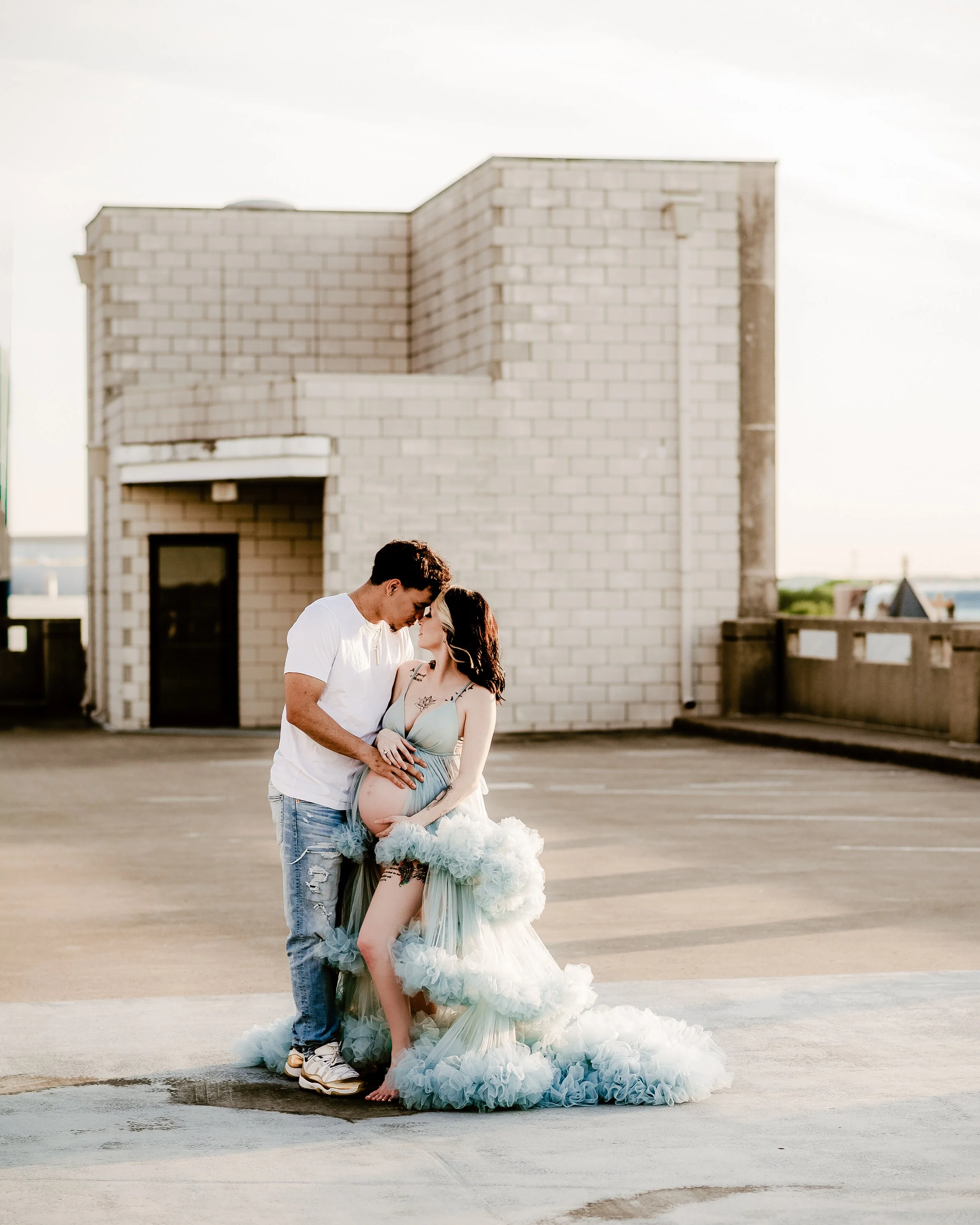 A couple, with the woman being pregnant, standing close together on a rooftop parking lot, with a brick building in the background. The woman is dressed in a flowing, ruffled gown, and the man in casual clothes. They are touching foreheads and lookin