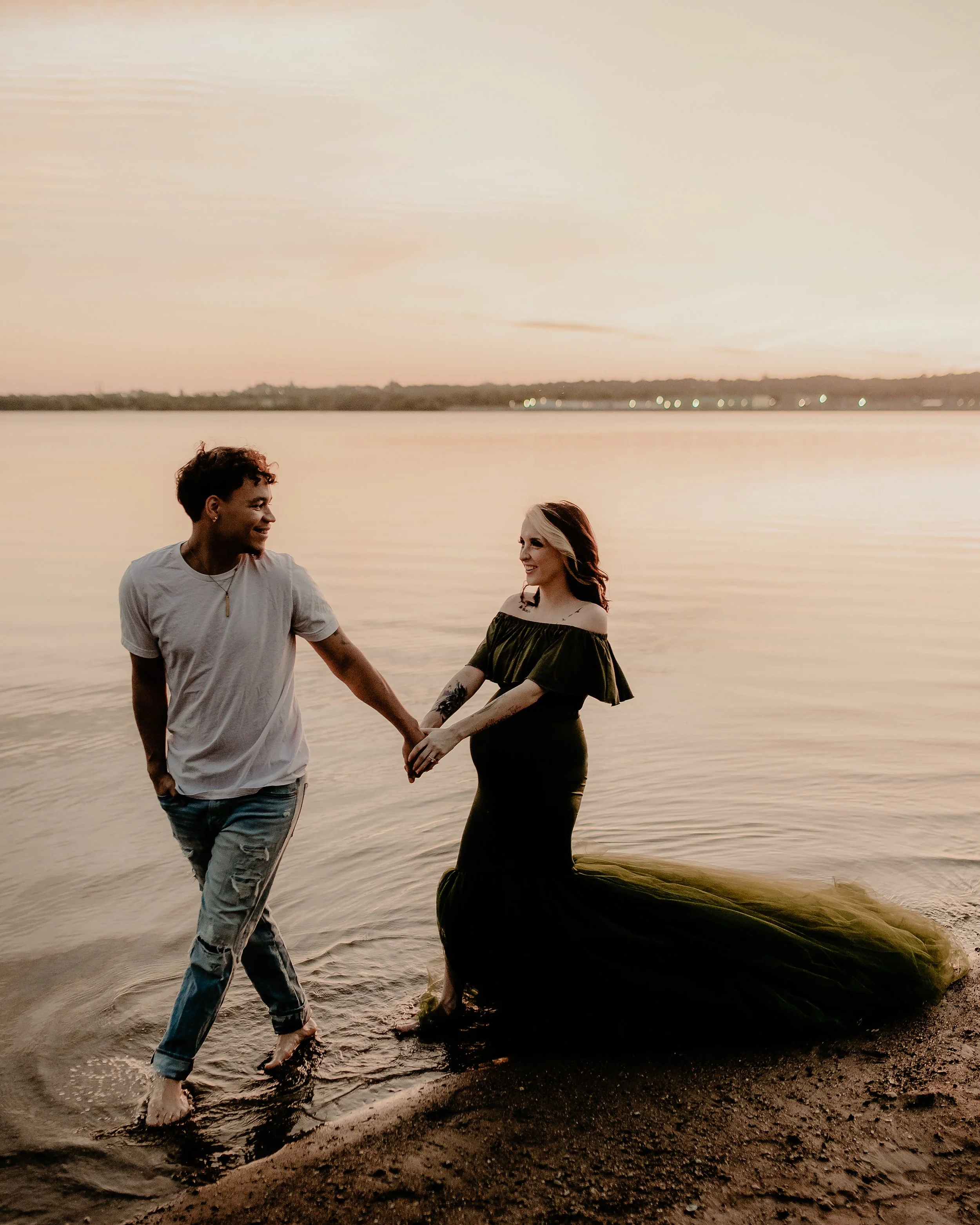 A man and a woman holding hands and walking into a lake at sunset. The woman is wearing an elegant off-the-shoulder green gown with a train, and the man is wearing a casual t-shirt and ripped jeans.