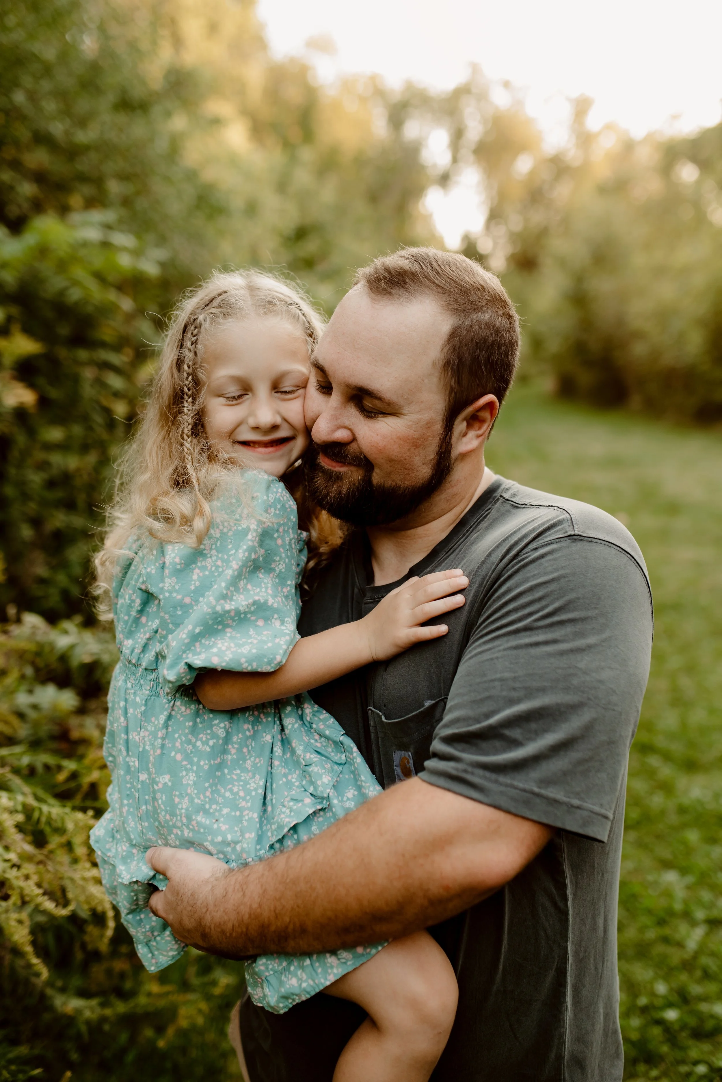 A man holding a young girl in a green outdoor setting at sunset, both smiling and touching faces affectionately.