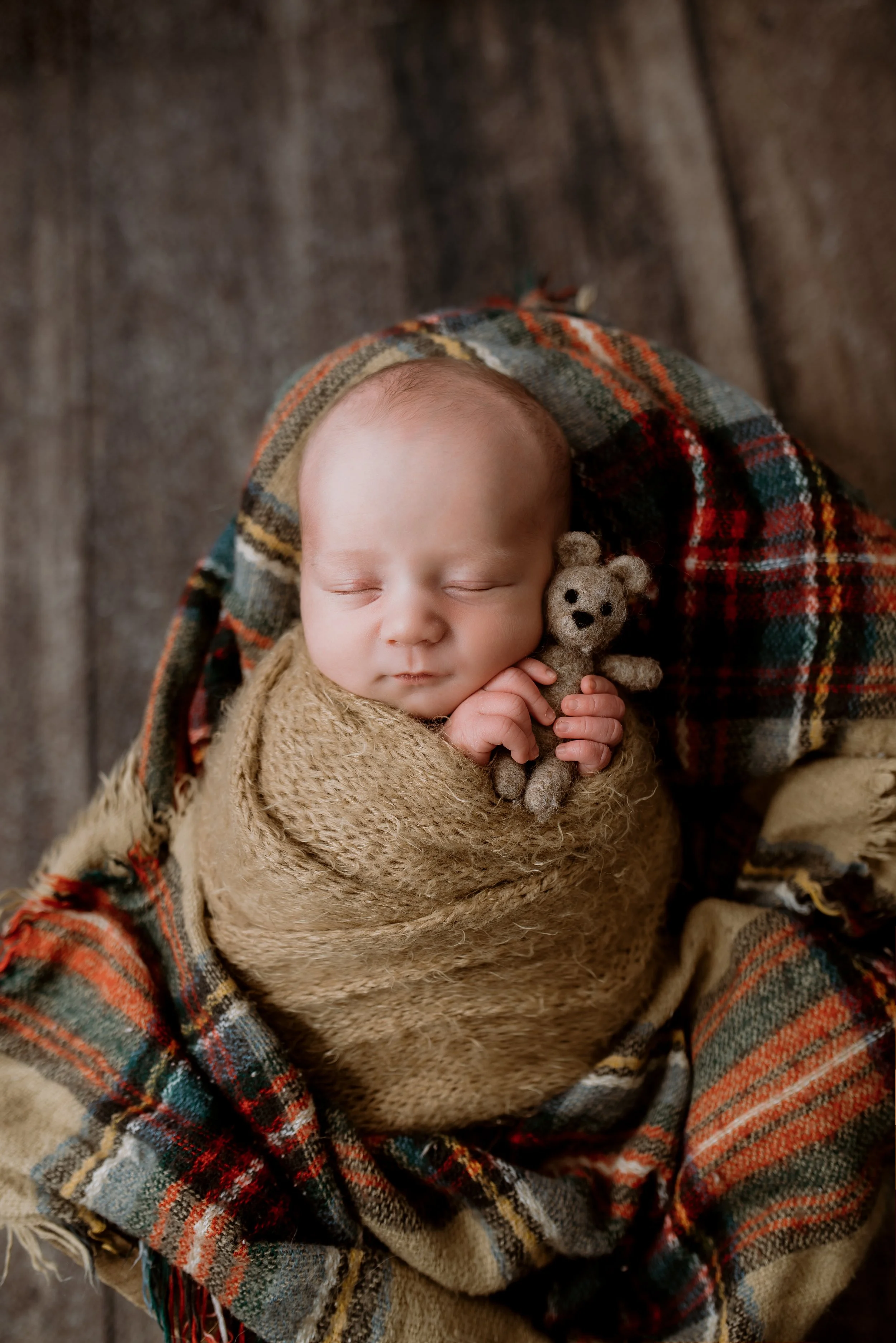 A sleeping baby wrapped in a textured beige shawl, holding a small felted teddy bear, with a plaid blanket draped around in warm tones, resting on a wooden surface.