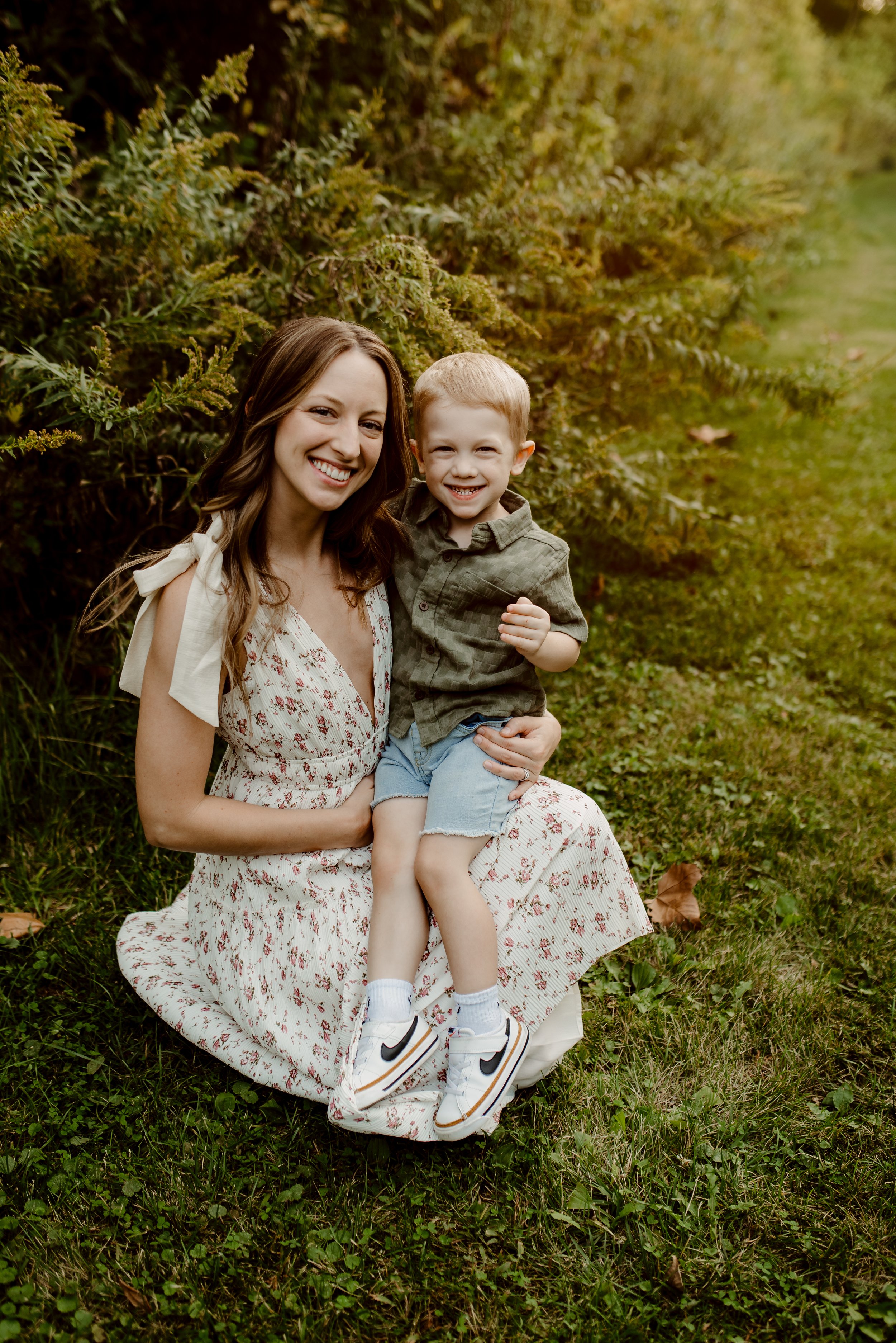 A smiling woman with long brown hair and a young boy sitting outdoors on grass, with greenery in the background, both happily posing for the camera.