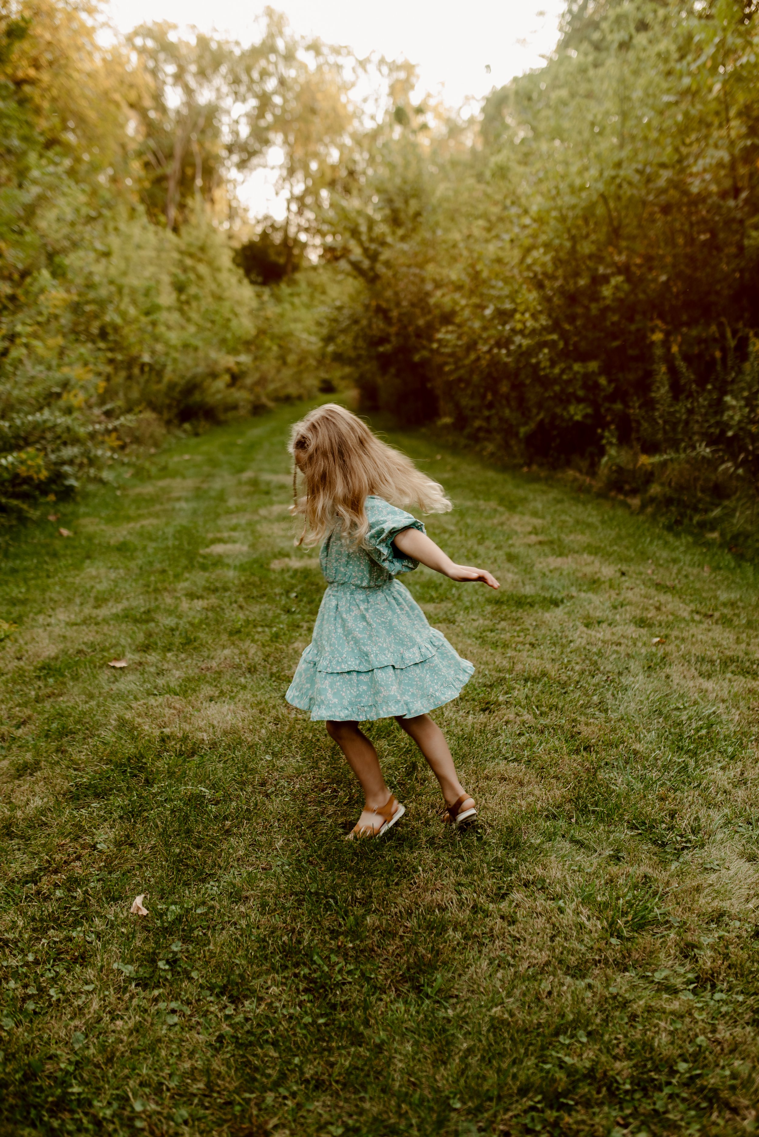 A young girl with long blonde hair wearing a light blue dress dancing on a grassy path surrounded by trees.