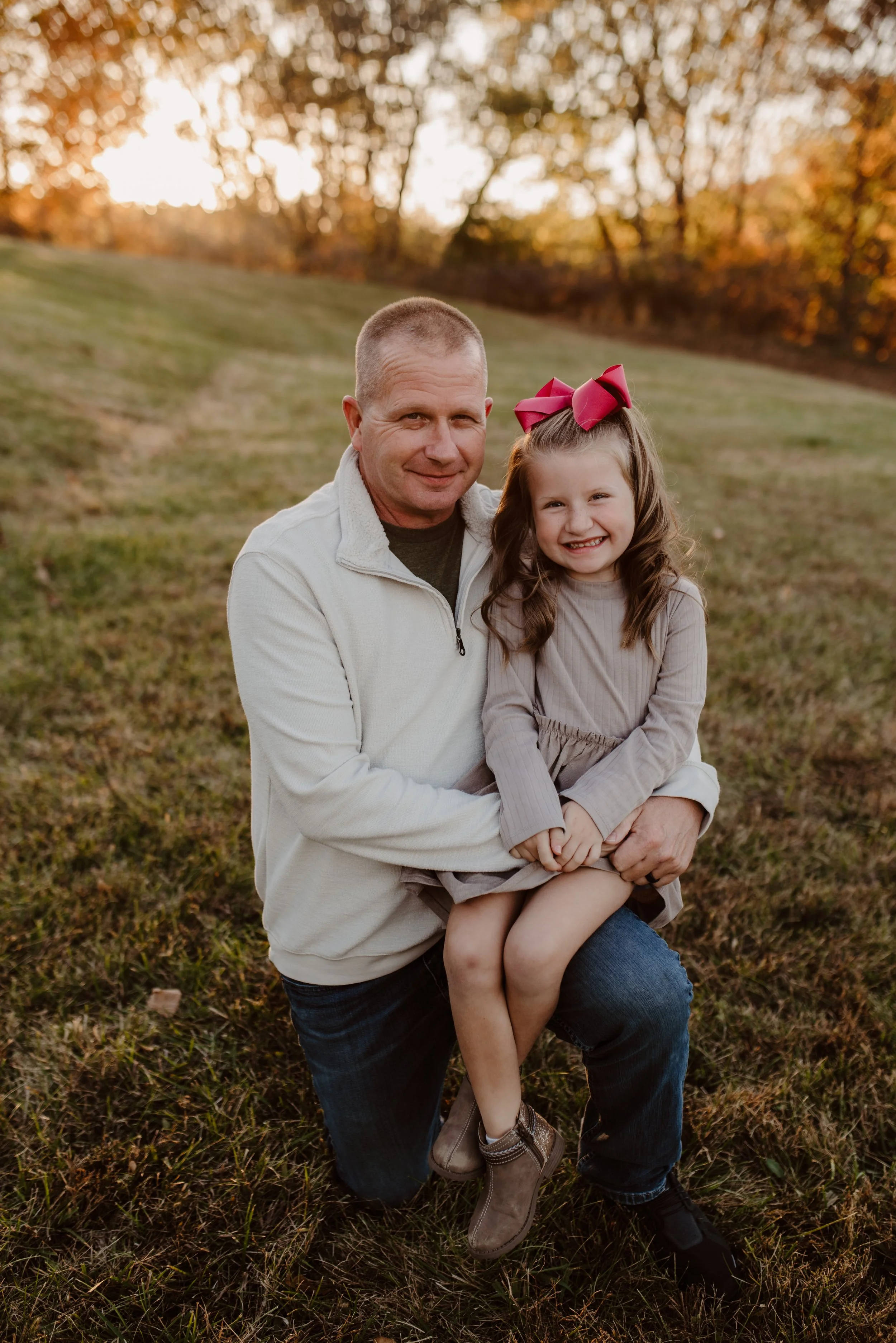 A man kneeling outdoors on grass, holding a young girl on his lap, both smiling at the camera during sunset with trees in the background.
