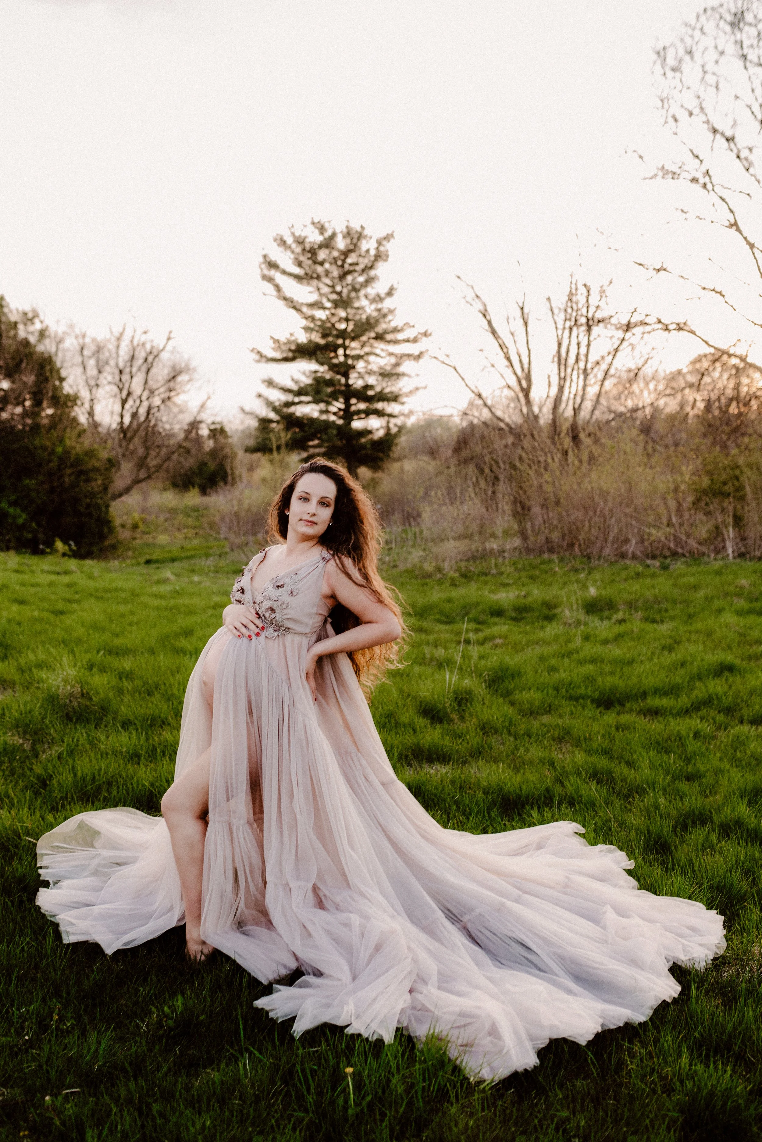 A pregnant woman in a flowing beige dress standing barefoot on a grassy field during sunset, with trees in the background.
