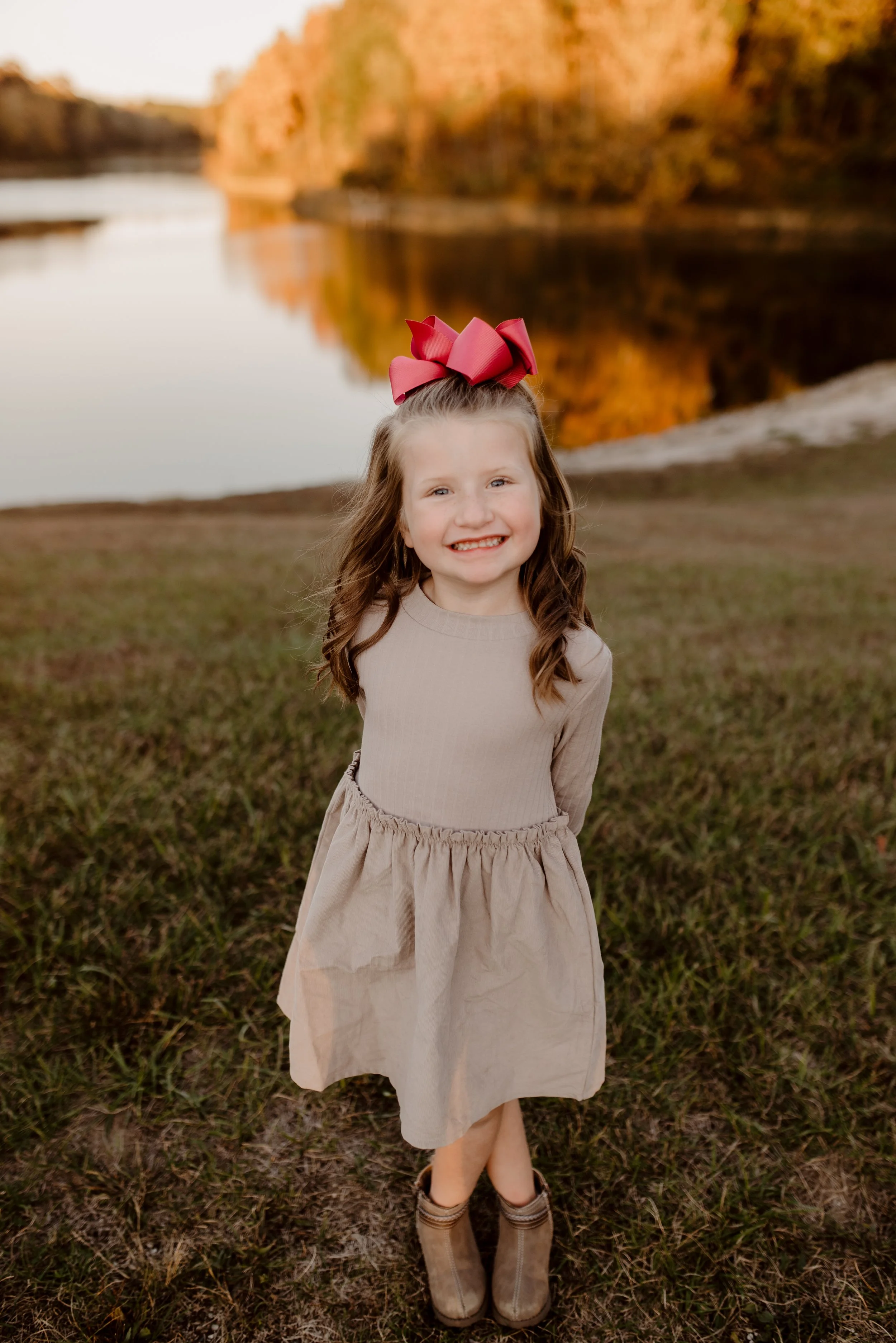 A young girl with long, wavy hair and a big pink bow on her head stands outdoors on a grassy field near a river during autumn sunset, smiling at the camera.