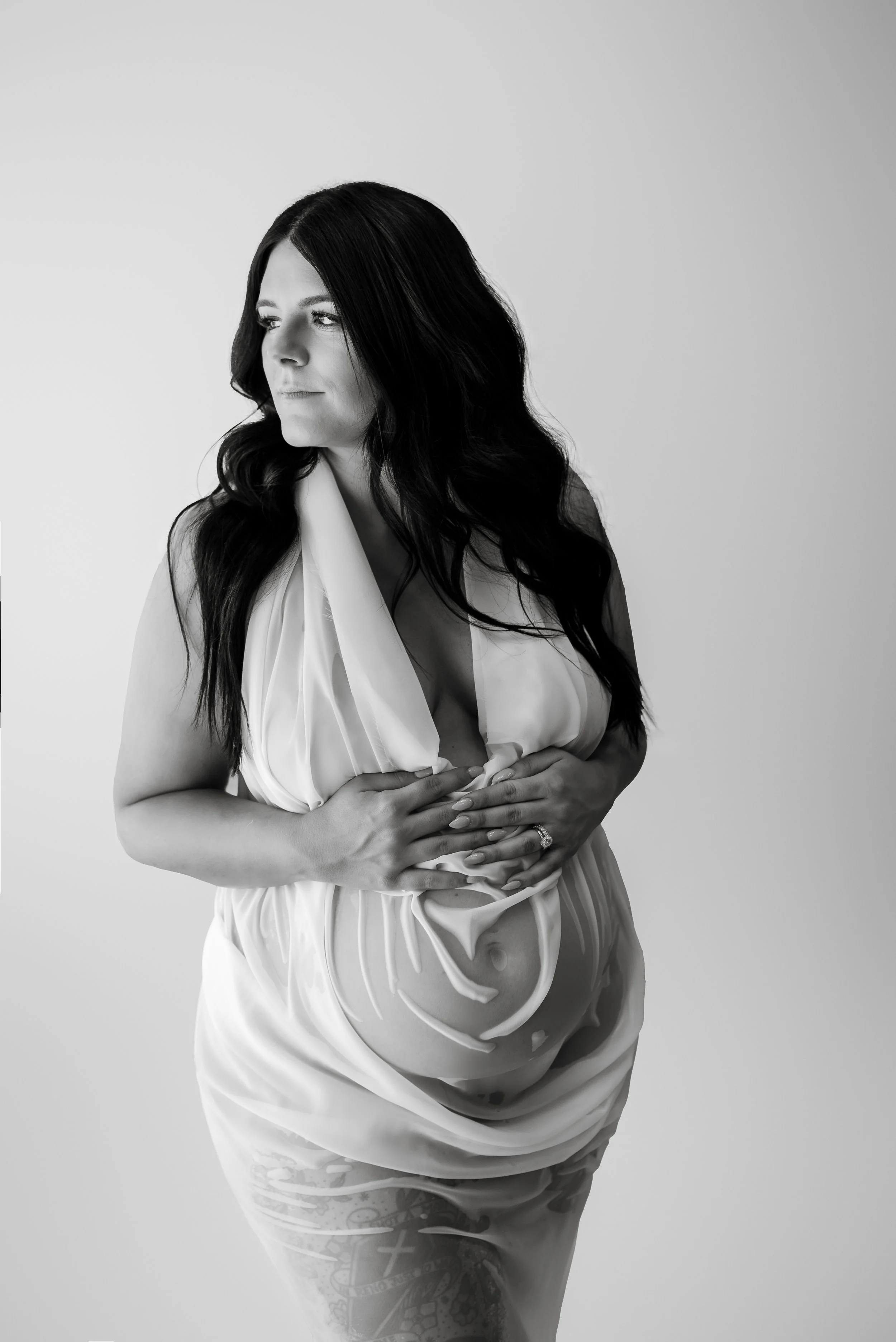 Black and white photo of a pregnant woman with long dark hair, wearing a draped, satin dress, gently holding her belly, standing against a plain background.