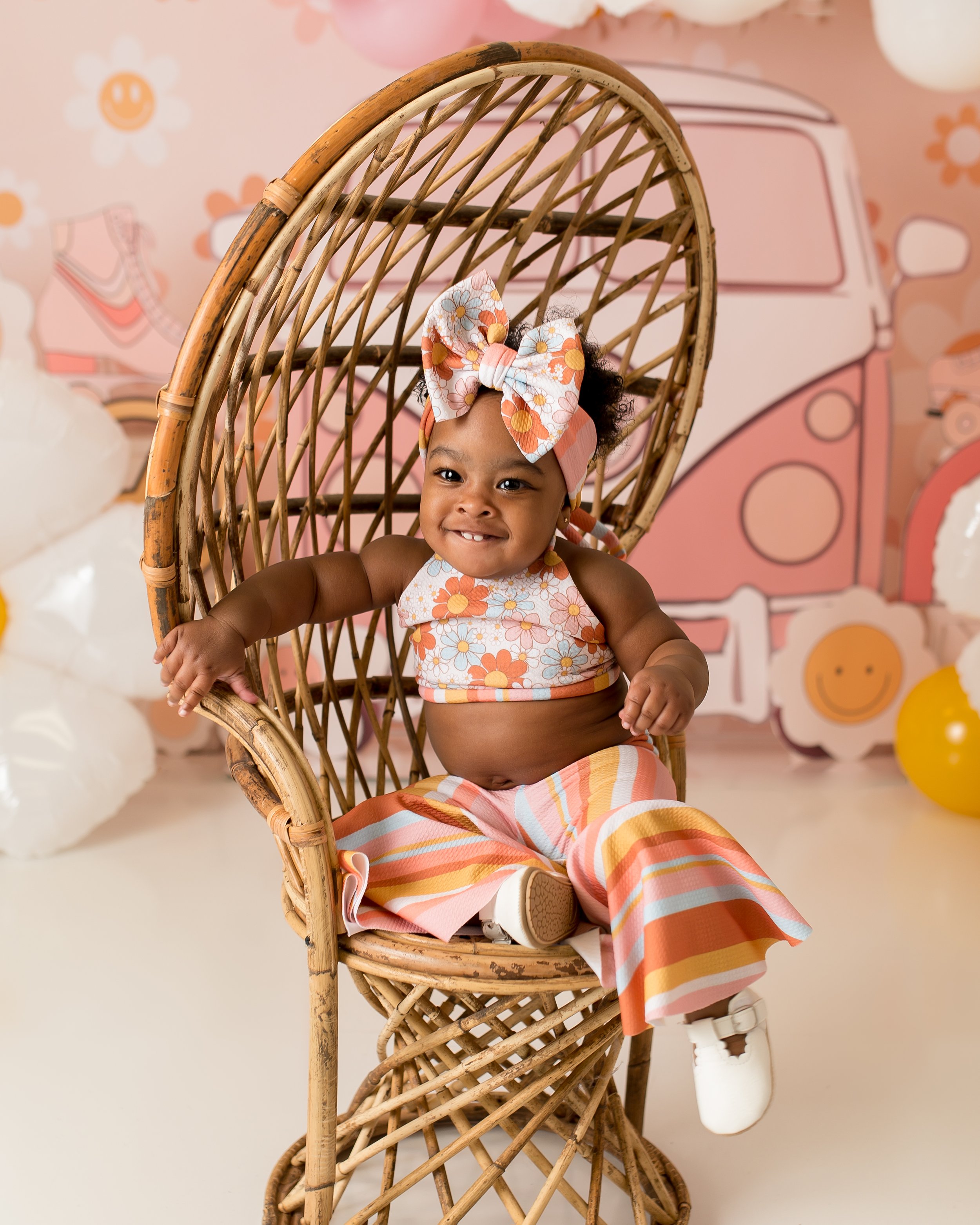 A smiling young girl sitting in a woven rattan chair with a floral outfit and matching headband, surrounded by a playful, colorful background featuring a pink van and smiley cloud decorations.