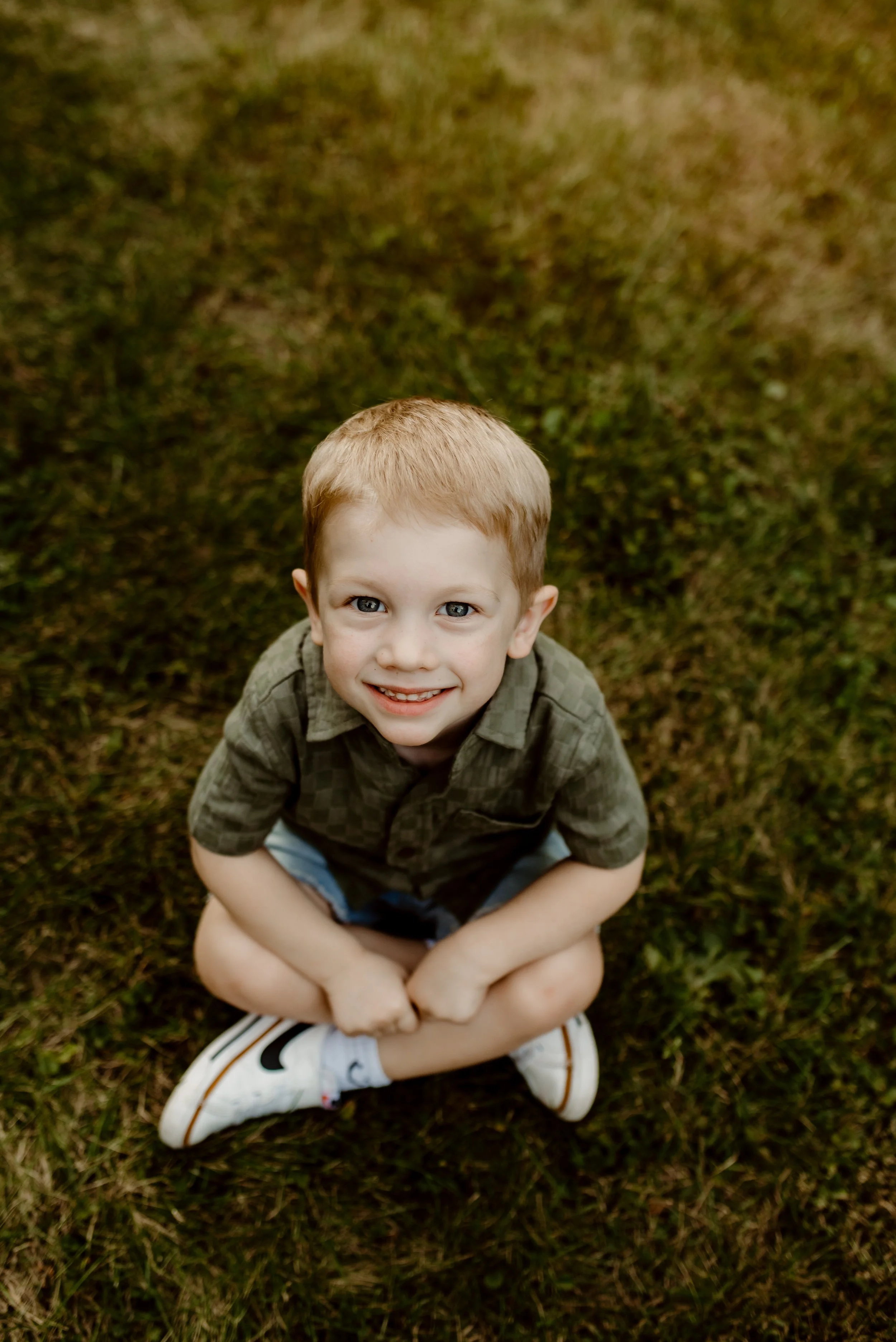 A young boy with red hair, blue eyes, and a big smile, sitting cross-legged on grass outdoors.