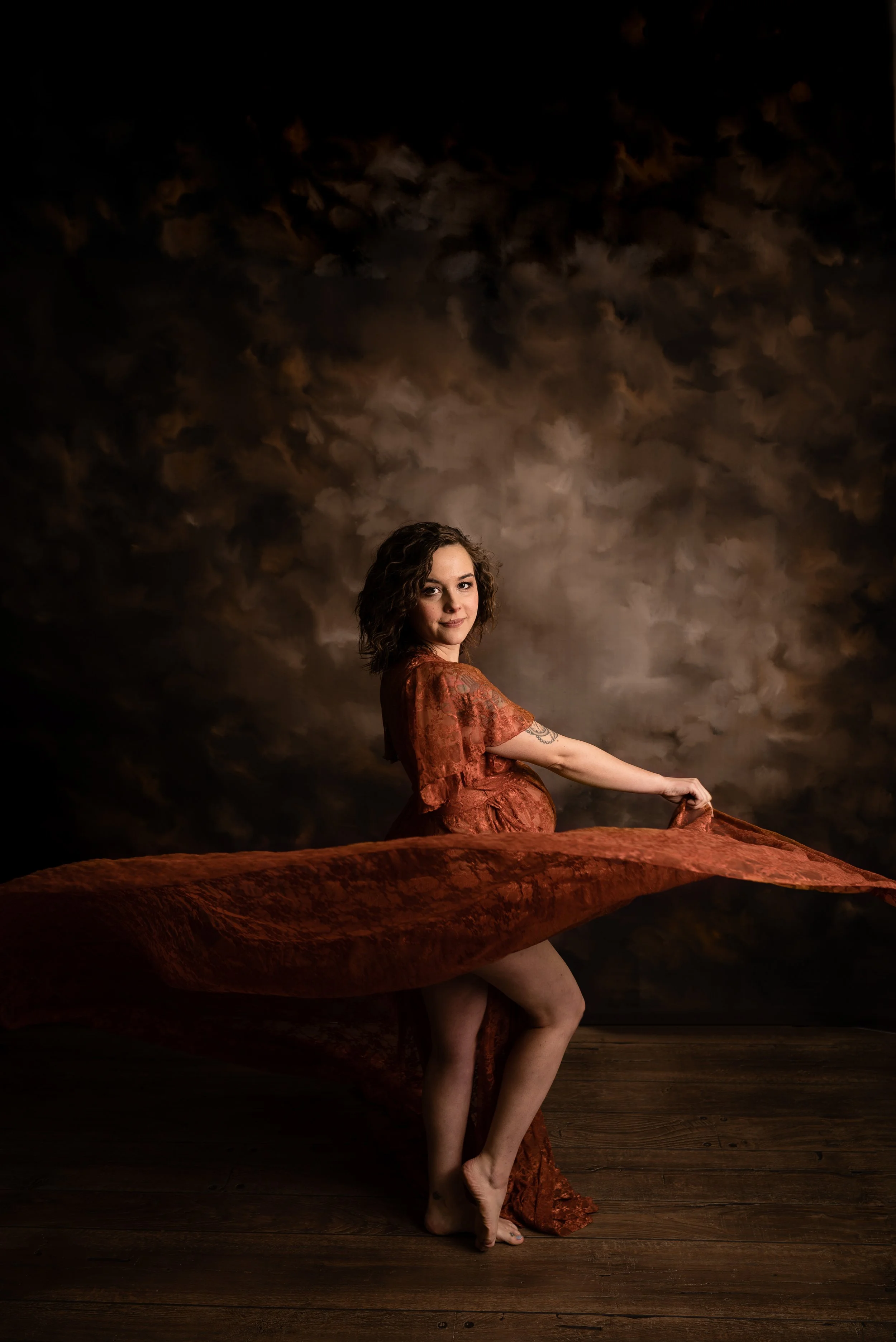A woman with curly hair in a reddish-orange dress standing barefoot on a wooden floor, holding the edge of her dress, with a dark, cloudy sky painted backdrop.