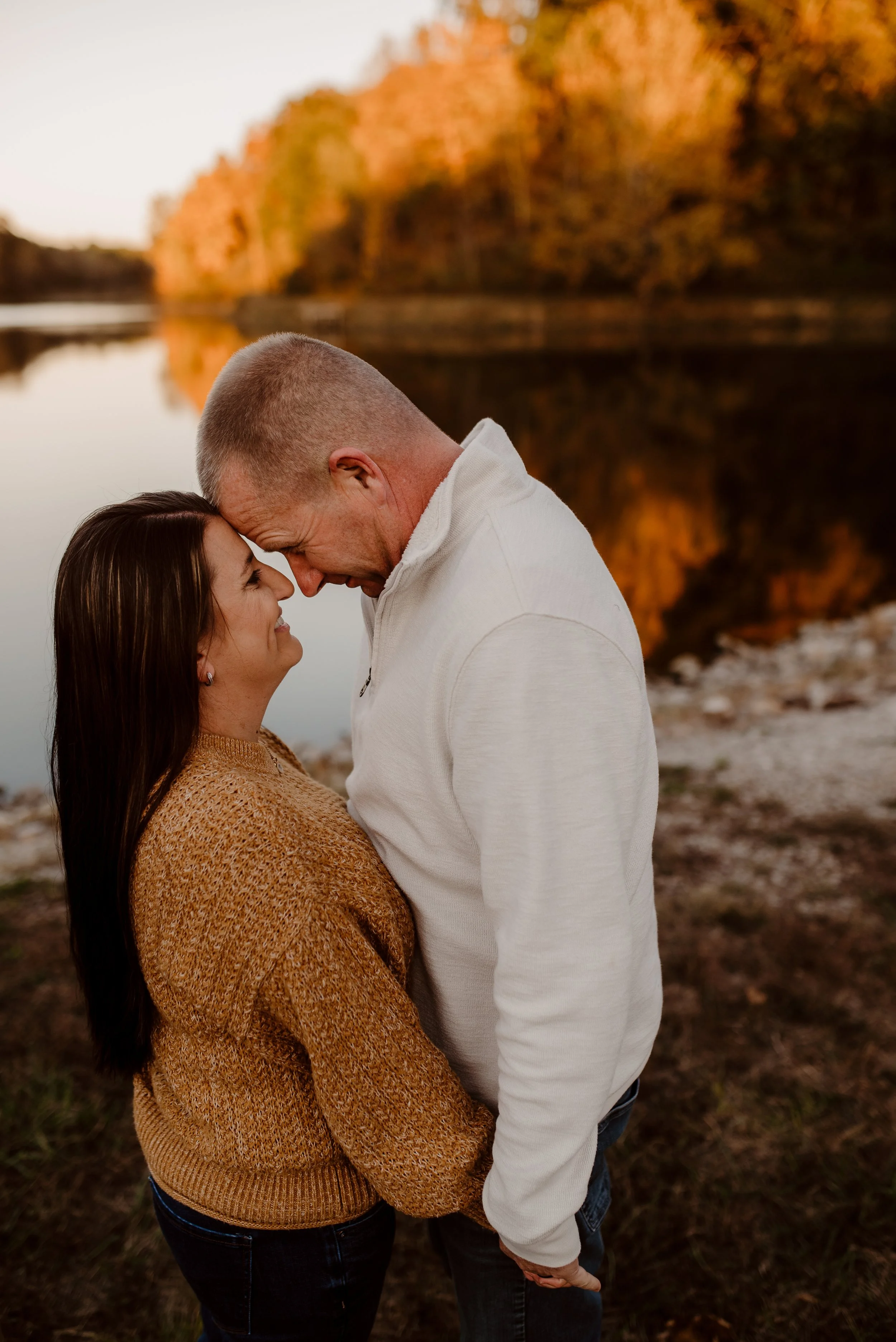 A couple standing close together by a lake with autumn trees in the background, smiling and touching foreheads during sunset.