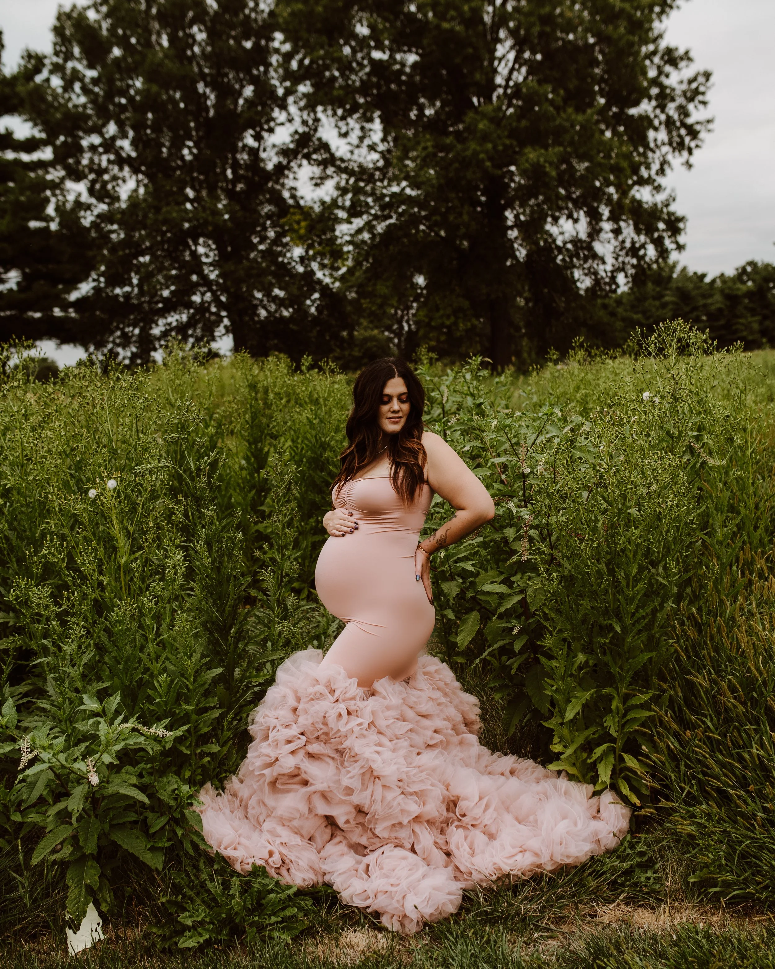A pregnant woman in a pale pink, ruffled gown standing in a field of tall green plants with large trees in the background.