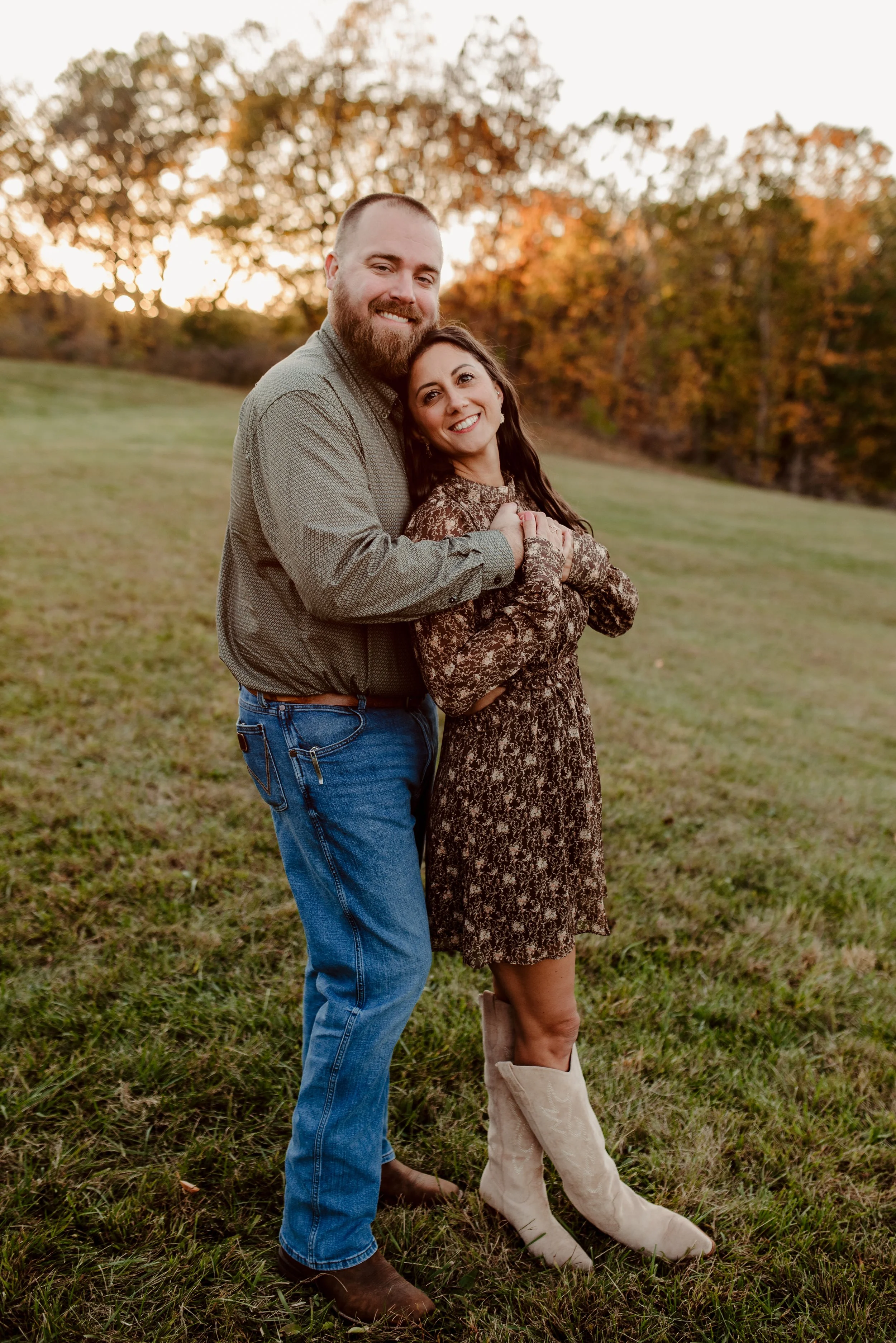 A happy couple standing in a grassy field during autumn sunset, embracing and smiling at the camera with colorful fall trees in the background.